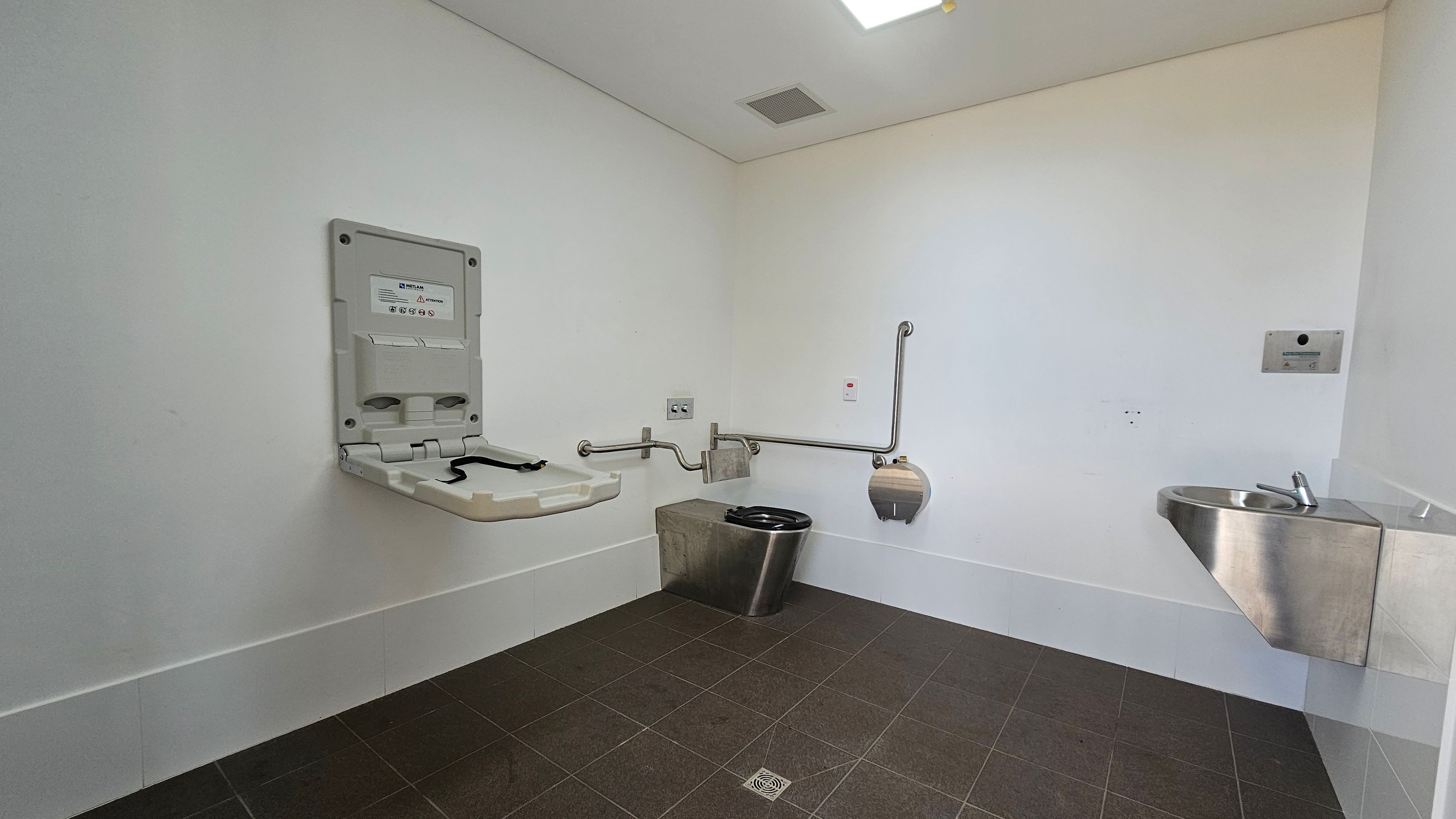 A public restroom with a baby changing station on the left wall. In the centre, there is a stainless steel toilet with grab bars on both sides and a toilet paper dispenser. On the right, a small stainless steel sink is mounted on the wall. The floor is tiled in dark grey, and the ceiling is plain white with a vent and a light fixture. A public restroom with a baby changing station on the left wall. In the centre, there is a stainless steel toilet with grab bars on both sides and a toilet paper dispenser. On the right, a small stainless steel sink is mounted on the wall. The floor is tiled in dark grey, and the ceiling is plain white with a vent and a light fixture.