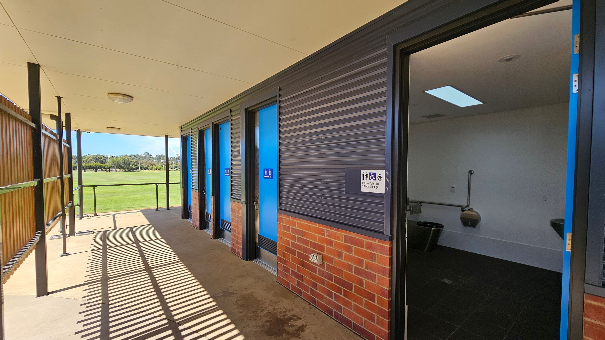 A row of public toilets with blue doors is on the right, set against a wall with dark corrugated panels and a brick base. One door is open, revealing a white interior with a toilet and grab bars. A sign indicates unisex, disabled, and baby change facilities. On the left, a wooden slatted fence casts shadows on the concrete floor. The ceiling is light-coloured with circular lights. In the background, a grassy field and trees are visible. A row of public toilets with blue doors is on the right, set against a wall with dark corrugated panels and a brick base. One door is open, revealing a white interior with a toilet and grab bars. A sign indicates unisex, disabled, and baby change facilities. On the left, a wooden slatted fence casts shadows on the concrete floor. The ceiling is light-coloured with circular lights. In the background, a grassy field and trees are visible.
