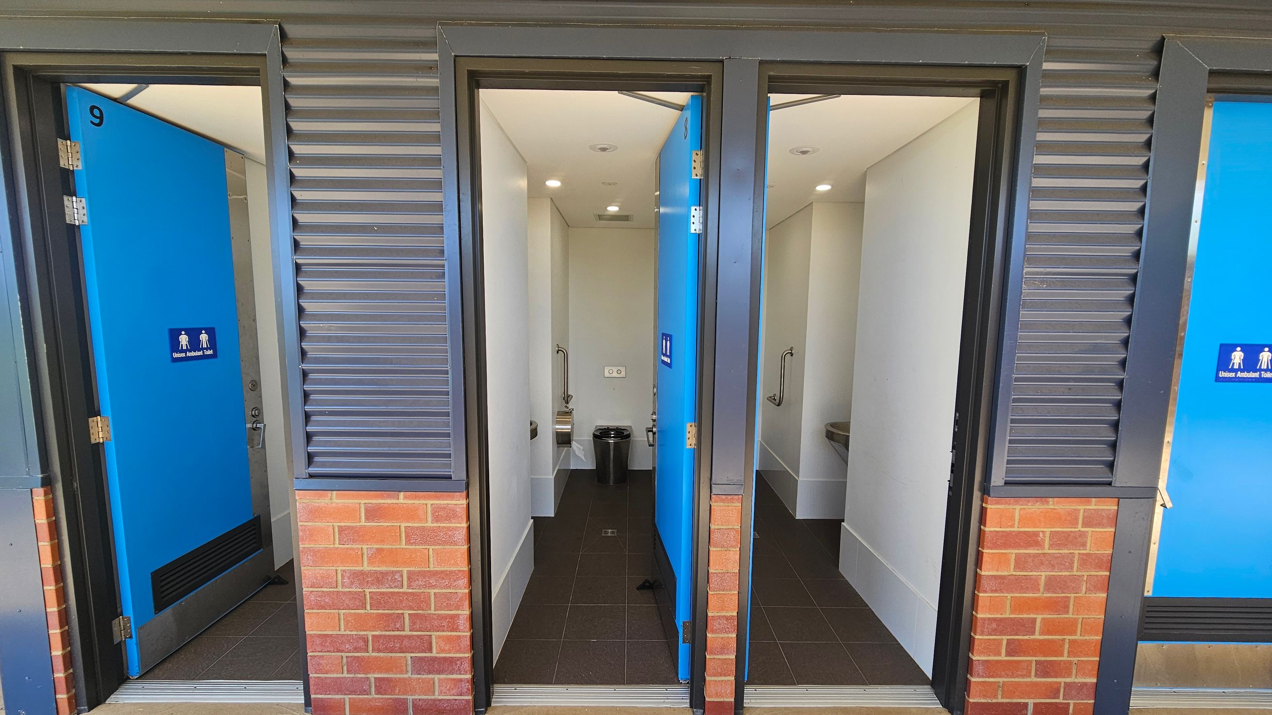 Three public toilet cubicles with blue doors, each labelled as unisex ambulant toilets. The doors are partially open, revealing white interiors with grab bars and waste bins. The exterior wall features a combination of grey metal cladding and red brick. The floor inside the cubicles is dark tiled, and the ceiling is white with recessed lighting. Three public toilet cubicles with blue doors, each labelled as unisex ambulant toilets. The doors are partially open, revealing white interiors with grab bars and waste bins. The exterior wall features a combination of grey metal cladding and red brick. The floor inside the cubicles is dark tiled, and the ceiling is white with recessed lighting.