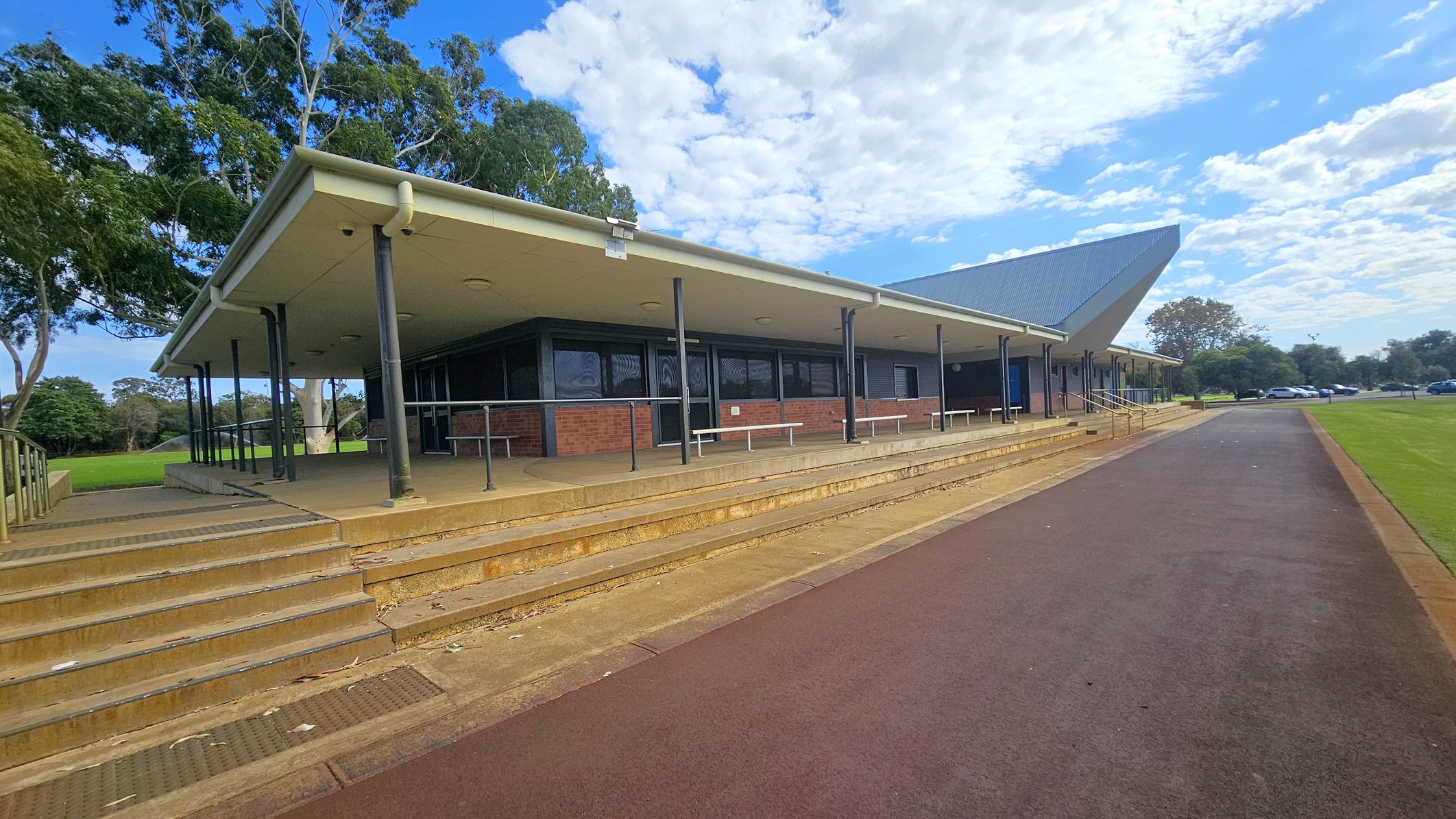 A building with a sloped roof and a covered walkway supported by black poles. The structure is made of brick with large windows. Steps lead up to the entrance, and there are benches along the walkway. On the left, there are trees with green foliage. On the right, a paved path runs alongside a grassy area, with parked cars visible in the distance. The sky is partly cloudy. A building with a sloped roof and a covered walkway supported by black poles. The structure is made of brick with large windows. Steps lead up to the entrance, and there are benches along the walkway. On the left, there are trees with green foliage. On the right, a paved path runs alongside a grassy area, with parked cars visible in the distance. The sky is partly cloudy.