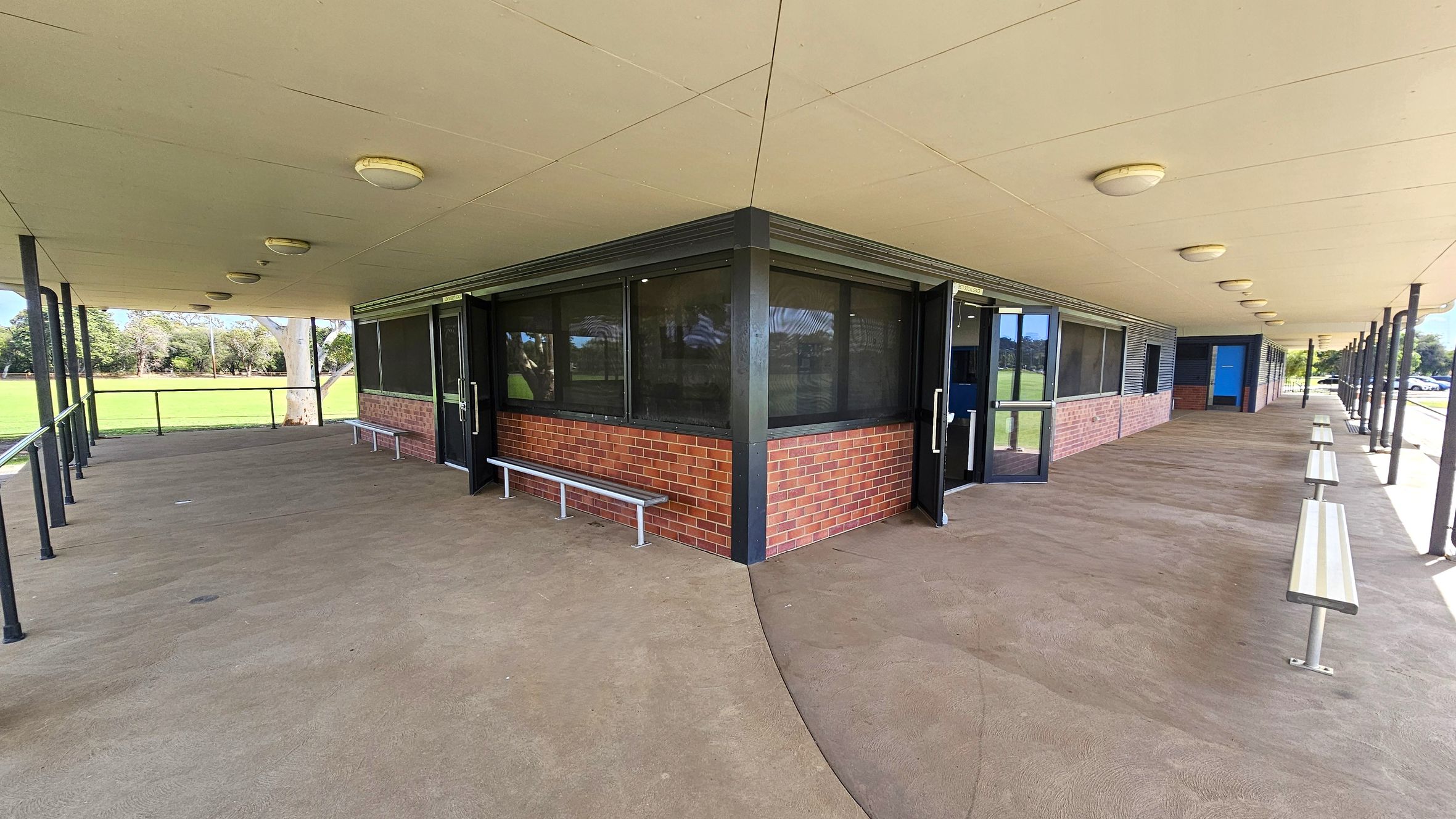 A covered outdoor area with a beige ceiling featuring round lights. The floor is concrete, and there are metal benches along the walls. The building has brick walls with large windows and glass doors. On the left, there's a grassy field with trees in the background. On the right, the structure extends with more benches and a blue door visible. The area is supported by black metal poles. A covered outdoor area with a beige ceiling featuring round lights. The floor is concrete, and there are metal benches along the walls. The building has brick walls with large windows and glass doors. On the left, there's a grassy field with trees in the background. On the right, the structure extends with more benches and a blue door visible. The area is supported by black metal poles.