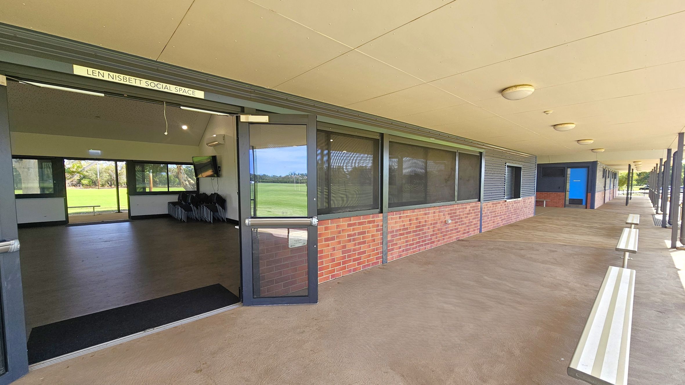 A covered outdoor walkway with a beige ceiling and concrete floor. On the left, a door is open to a room labelled "Len Nisbett Social Space," revealing a dark floor and windows overlooking a grassy field. The right side features a brick and metal-panelled wall with several windows and a blue door. Benches are positioned along the walkway, and round ceiling lights are evenly spaced above. A covered outdoor walkway with a beige ceiling and concrete floor. On the left, a door is open to a room labelled "Len Nisbett Social Space," revealing a dark floor and windows overlooking a grassy field. The right side features a brick and metal-panelled wall with several windows and a blue door. Benches are positioned along the walkway, and round ceiling lights are evenly spaced above.