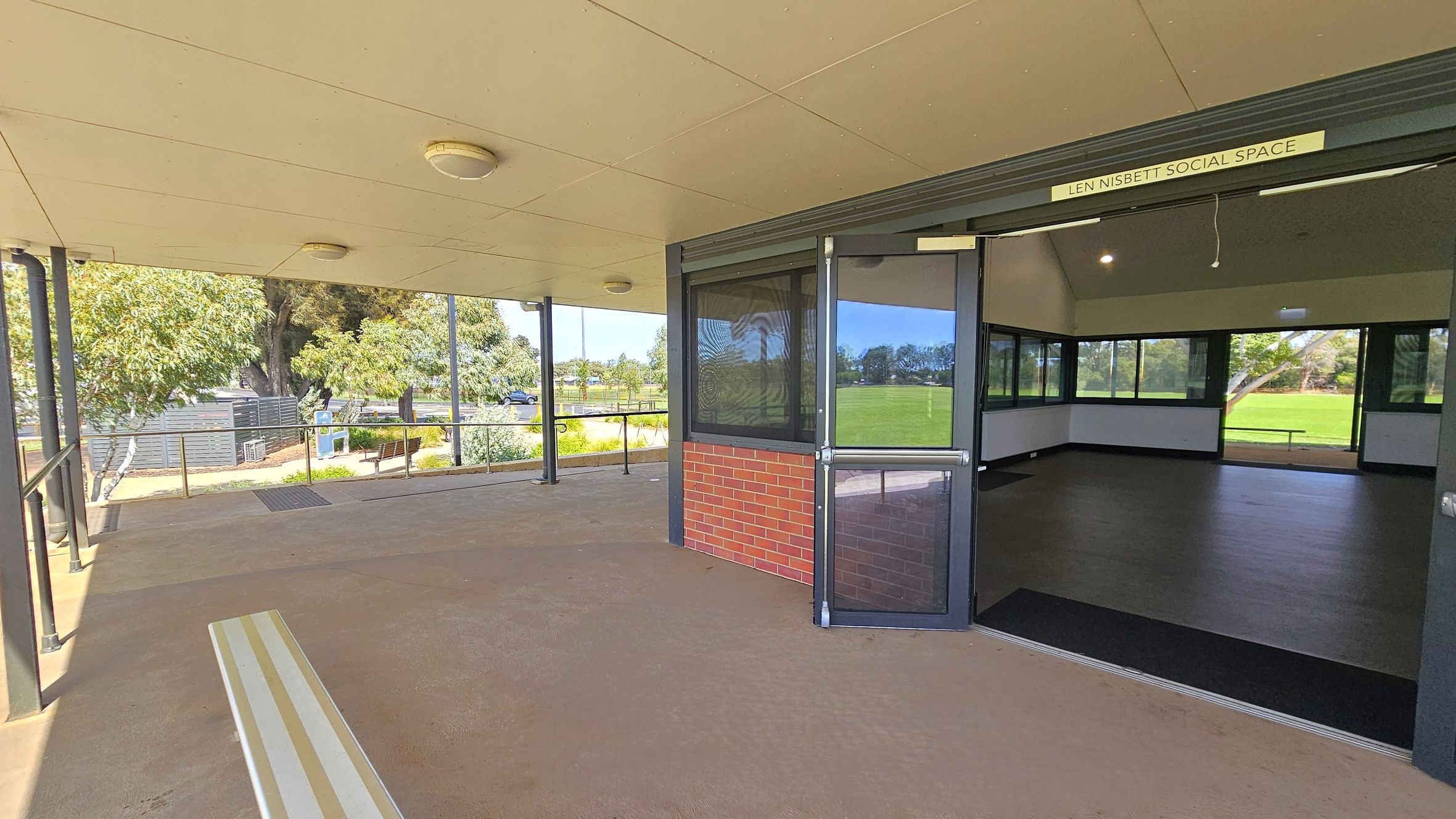 A covered outdoor area with a beige ceiling and several round lights. On the left, there are trees and a pathway with benches. The entrance to the "Len Nisbett Social Space" is on the right, featuring a glass door and brick wall. Inside, the room has dark flooring and large windows overlooking a grassy field. The floor outside is a light brown concrete. A covered outdoor area with a beige ceiling and several round lights. On the left, there are trees and a pathway with benches. The entrance to the "Len Nisbett Social Space" is on the right, featuring a glass door and brick wall. Inside, the room has dark flooring and large windows overlooking a grassy field. The floor outside is a light brown concrete.