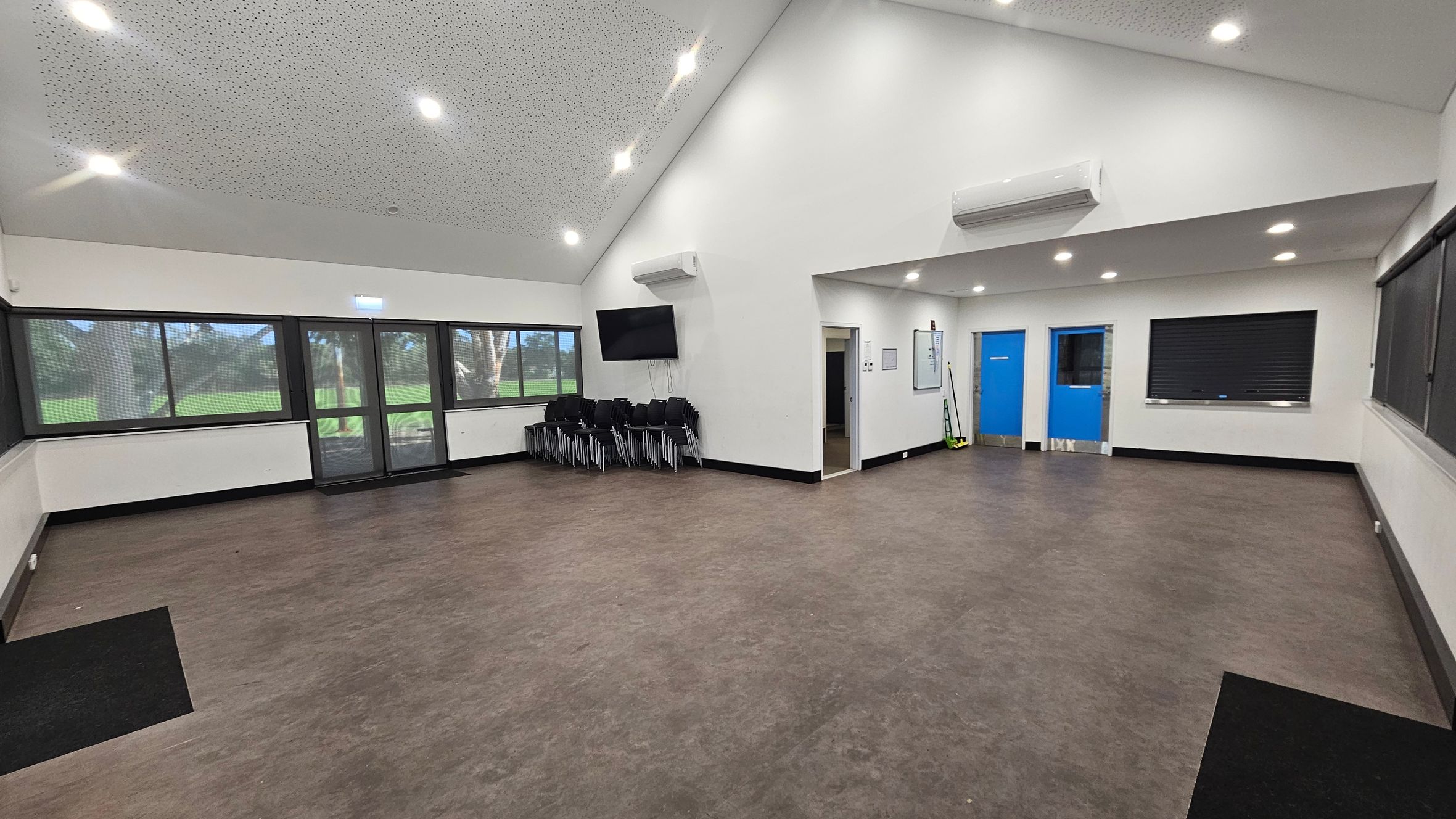 A spacious room with a brown floor and a white, perforated ceiling with recessed lights. On the left, large windows and a glass door offer a view of greenery outside. In the centre, a stack of black chairs is placed against the wall beneath a mounted TV. On the right, two bright blue doors are visible, along with a closed shutter window. A whiteboard and cleaning tools are mounted on the wall near the doors. The room has a clean and organised appearance. A spacious room with a brown floor and a white, perforated ceiling with recessed lights. On the left, large windows and a glass door offer a view of greenery outside. In the centre, a stack of black chairs is placed against the wall beneath a mounted TV. On the right, two bright blue doors are visible, along with a closed shutter window. A whiteboard and cleaning tools are mounted on the wall near the doors. The room has a clean and organised appearance.
