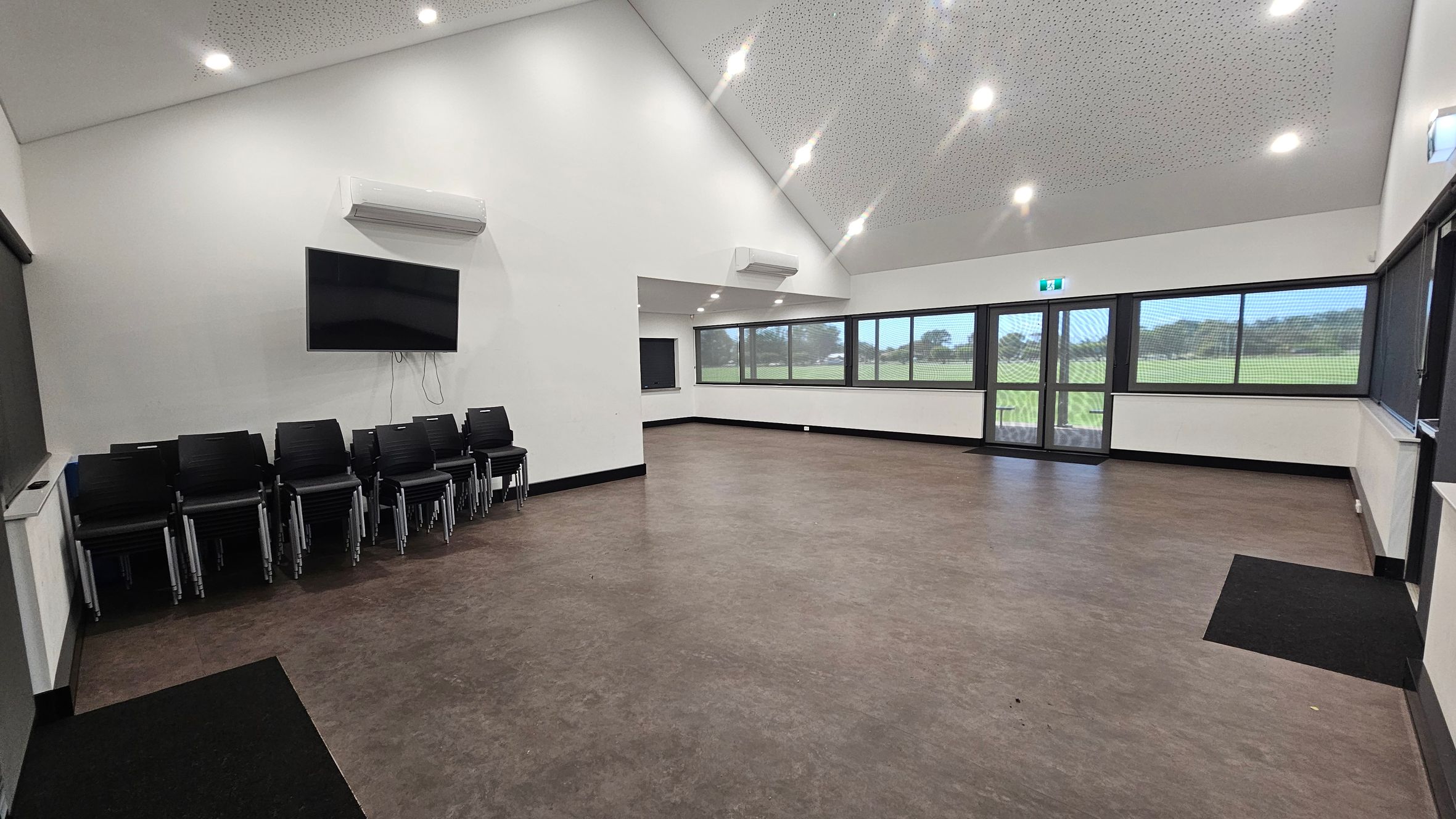 A spacious room with a high, sloped ceiling featuring recessed lights. On the left, there is a stack of black chairs beneath a wall-mounted television and an air conditioning unit. The floor is a mix of brown and purple tones. The right side has large windows and a glass door, offering a view of a grassy field outside. The walls are white, and there is another air conditioning unit above the windows. A spacious room with a high, sloped ceiling featuring recessed lights. On the left, there is a stack of black chairs beneath a wall-mounted television and an air conditioning unit. The floor is a mix of brown and purple tones. The right side has large windows and a glass door, offering a view of a grassy field outside. The walls are white, and there is another air conditioning unit above the windows.