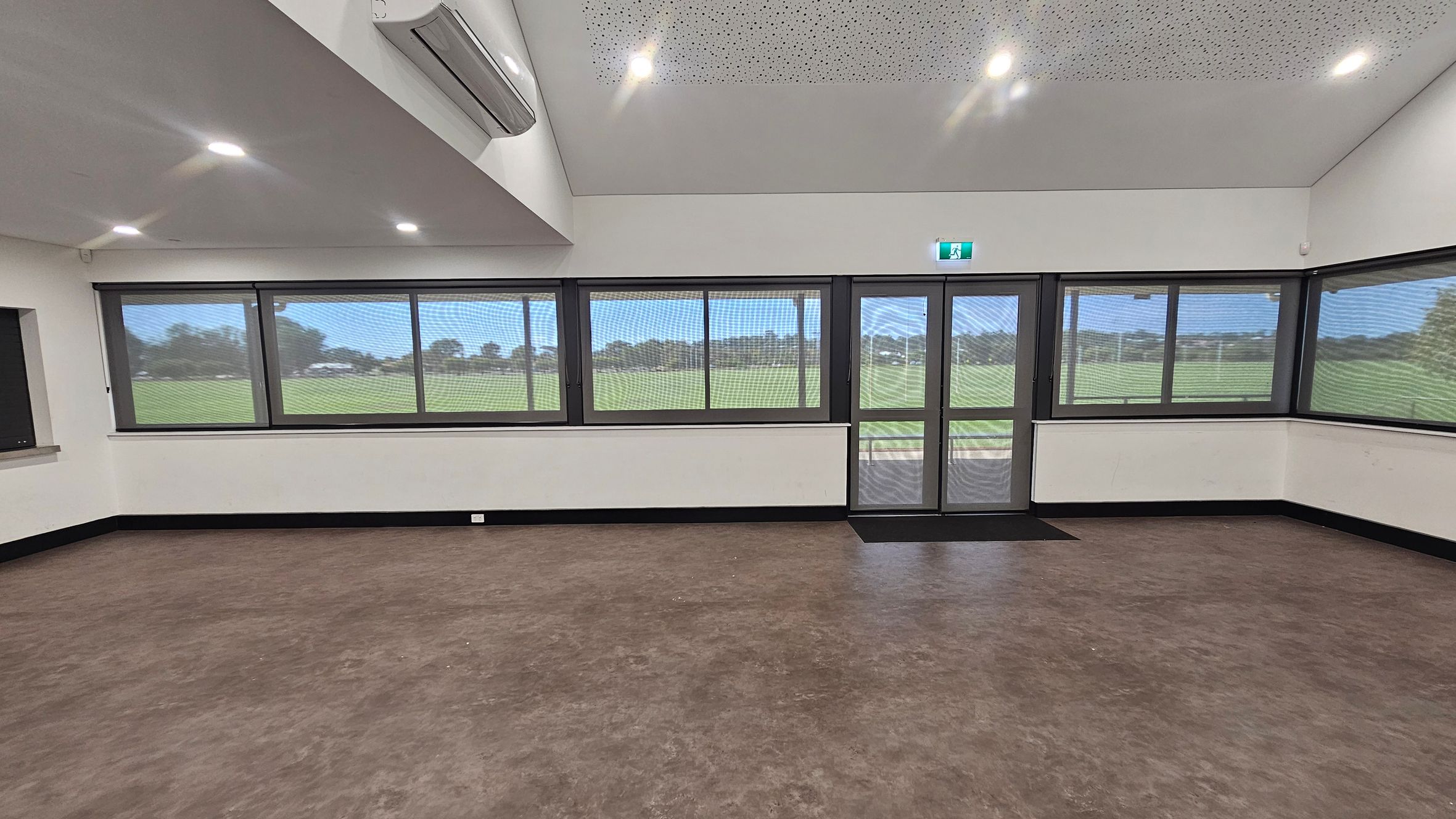 A room with large windows and a glass door on the right, offering a view of a grassy field outside. The walls are white, and the floor is brown with a smooth finish. The ceiling is white with recessed lighting and an air conditioning unit on the left. The windows have a mesh screen, and there is a black skirting board along the bottom of the walls. An emergency exit sign is above the door. A room with large windows and a glass door on the right, offering a view of a grassy field outside. The walls are white, and the floor is brown with a smooth finish. The ceiling is white with recessed lighting and an air conditioning unit on the left. The windows have a mesh screen, and there is a black skirting board along the bottom of the walls. An emergency exit sign is above the door.