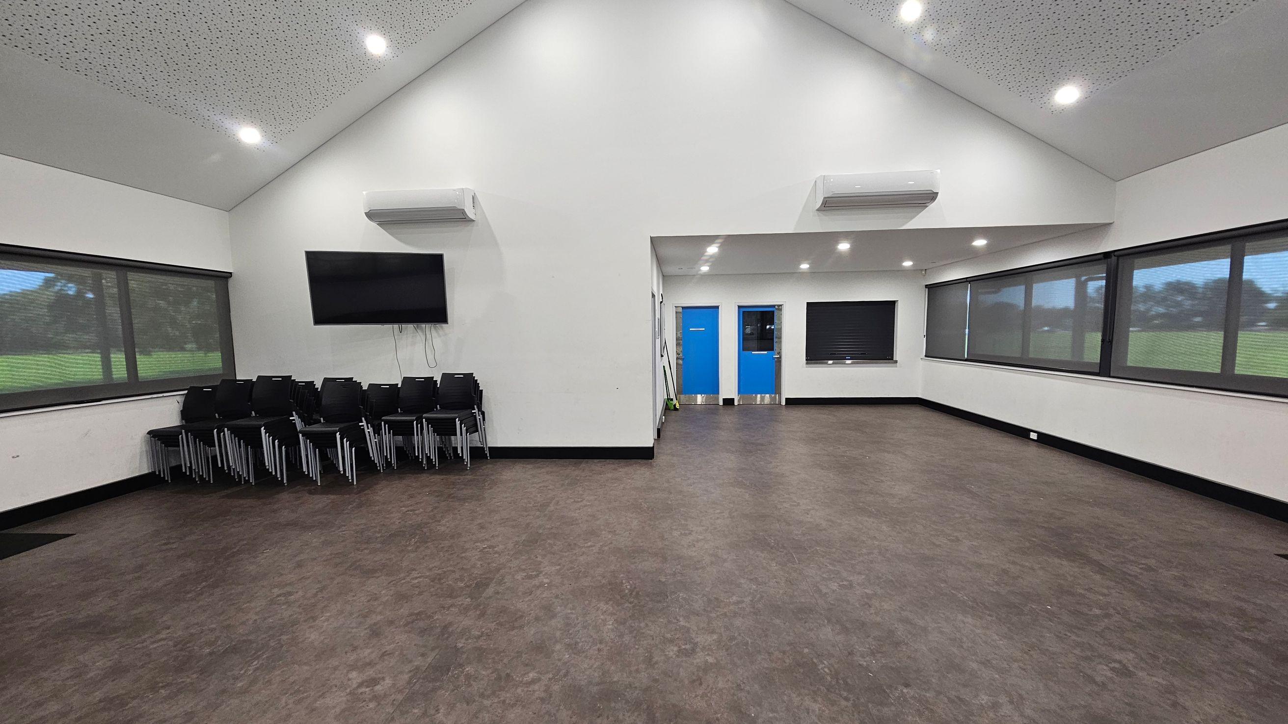 A spacious room with a high, sloped ceiling featuring recessed lights. On the left, there is a stack of black chairs beneath a wall-mounted television. The right side has large windows with blinds, offering a view of a grassy area outside. In the centre, two blue doors are visible, with a small window between them. The floor is a mix of brown and purple tones, and there are air conditioning units mounted on the walls. A spacious room with a high, sloped ceiling featuring recessed lights. On the left, there is a stack of black chairs beneath a wall-mounted television. The right side has large windows with blinds, offering a view of a grassy area outside. In the centre, two blue doors are visible, with a small window between them. The floor is a mix of brown and purple tones, and there are air conditioning units mounted on the walls.