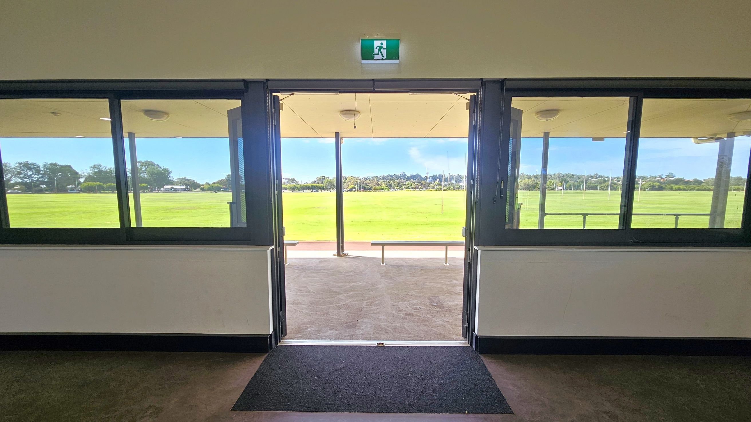 A view from inside a building looking out through a glass door. On either side of the door are large windows. Above the door is an emergency exit sign. Outside, there is a covered area with a bench and a wide expanse of green grass, possibly a sports field. Trees and a few structures are visible in the distance. The floor inside is carpeted near the entrance, transitioning to a hard surface outside. The ceiling inside is plain with a light fixture. A view from inside a building looking out through a glass door. On either side of the door are large windows. Above the door is an emergency exit sign. Outside, there is a covered area with a bench and a wide expanse of green grass, possibly a sports field. Trees and a few structures are visible in the distance. The floor inside is carpeted near the entrance, transitioning to a hard surface outside. The ceiling inside is plain with a light fixture.