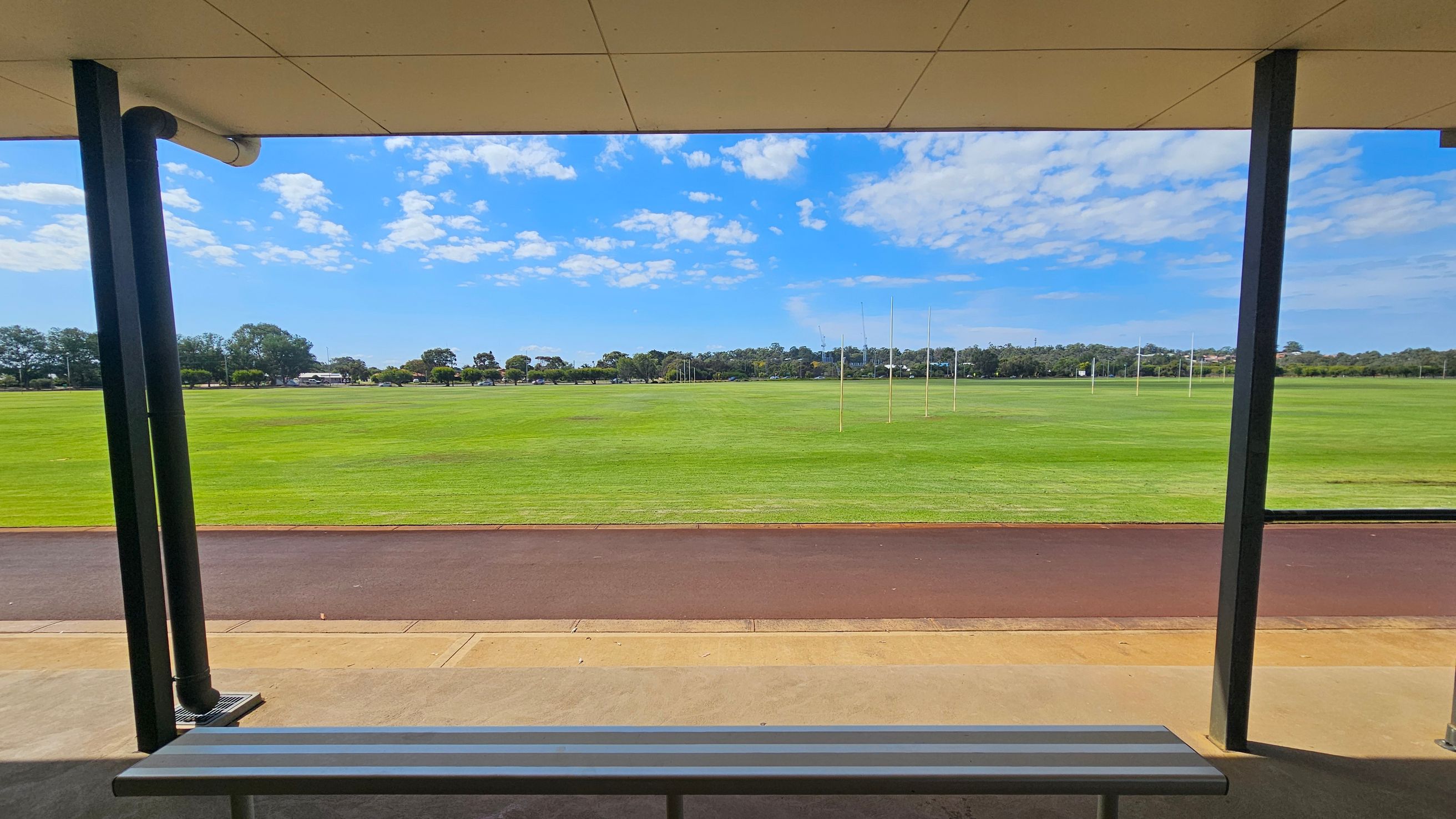 A sports field with green grass and several goalposts in the distance. In the foreground, a sheltered area with a metal bench is visible, supported by dark vertical poles. The ground is a mix of concrete and a reddish-brown track. Trees and a few buildings are visible on the horizon. The ceiling of the shelter is light-coloured and flat. A sports field with green grass and several goalposts in the distance. In the foreground, a sheltered area with a metal bench is visible, supported by dark vertical poles. The ground is a mix of concrete and a reddish-brown track. Trees and a few buildings are visible on the horizon. The ceiling of the shelter is light-coloured and flat.