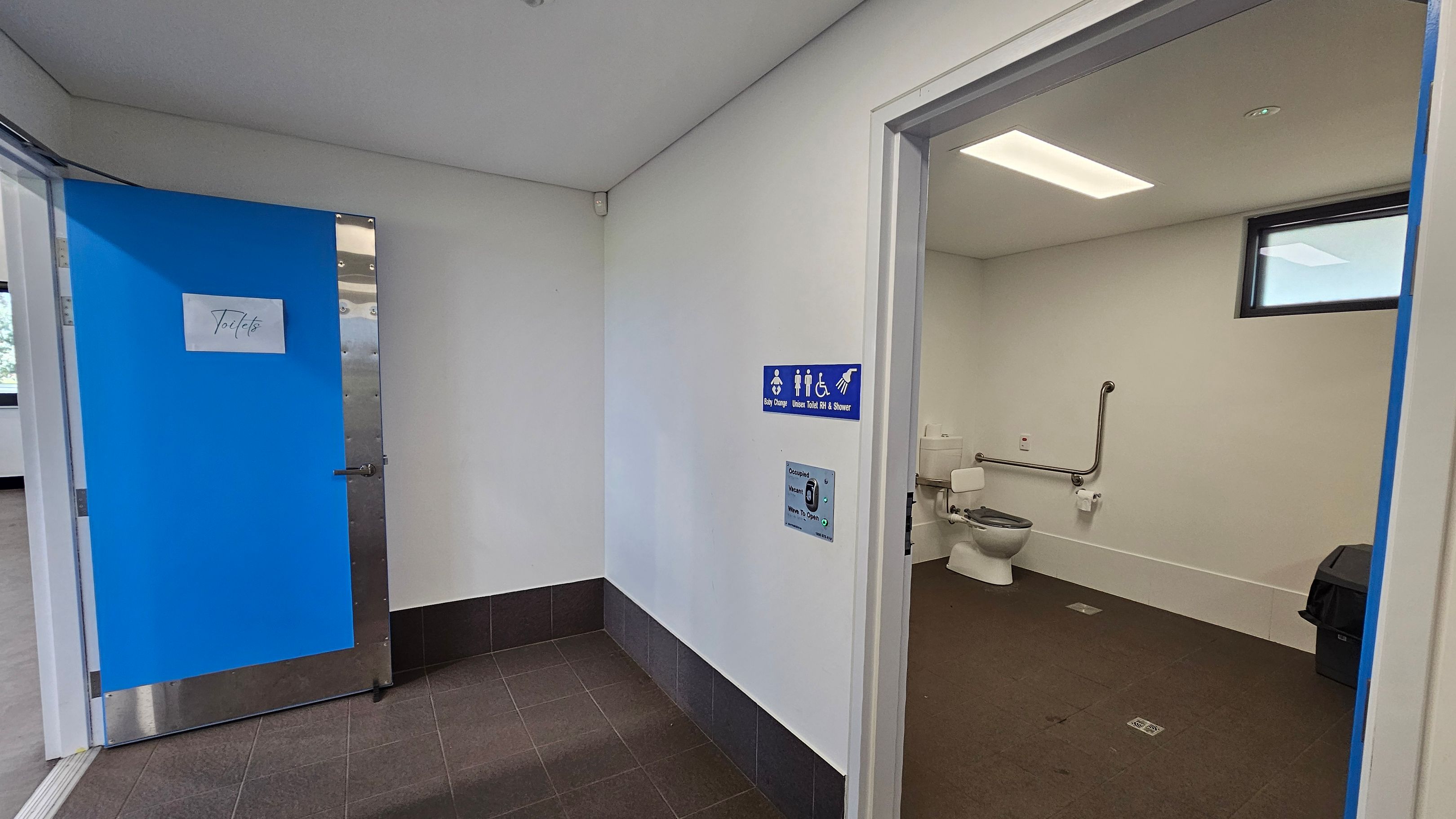 A public toilet area with a blue door on the left, labelled "Toilets". The floor is tiled in dark grey. On the right, an open door reveals an accessible bathroom with a toilet, grab bars, and a waste bin. A blue sign indicates facilities for baby change, toilets, and a shower. The ceiling is white with a rectangular light fixture, and a small window is visible in the bathroom. A public toilet area with a blue door on the left, labelled "Toilets". The floor is tiled in dark grey. On the right, an open door reveals an accessible bathroom with a toilet, grab bars, and a waste bin. A blue sign indicates facilities for baby change, toilets, and a shower. The ceiling is white with a rectangular light fixture, and a small window is visible in the bathroom.