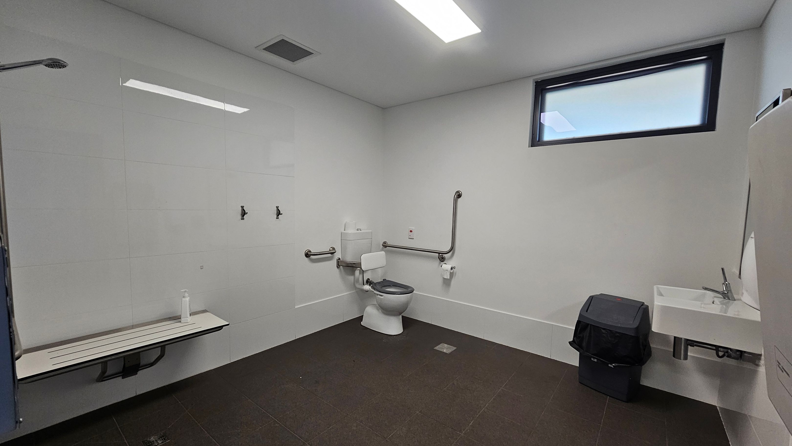 A spacious, accessible bathroom with white tiled walls and a dark tiled floor. On the left, a foldable bench with a bottle on it. In the centre, a toilet with support rails on both sides and a toilet paper holder. Above the toilet, a small window provides natural light. On the right, a white sink with a tap, and a black bin underneath. The ceiling has a rectangular light fixture and a vent. A spacious, accessible bathroom with white tiled walls and a dark tiled floor. On the left, a foldable bench with a bottle on it. In the centre, a toilet with support rails on both sides and a toilet paper holder. Above the toilet, a small window provides natural light. On the right, a white sink with a tap, and a black bin underneath. The ceiling has a rectangular light fixture and a vent.