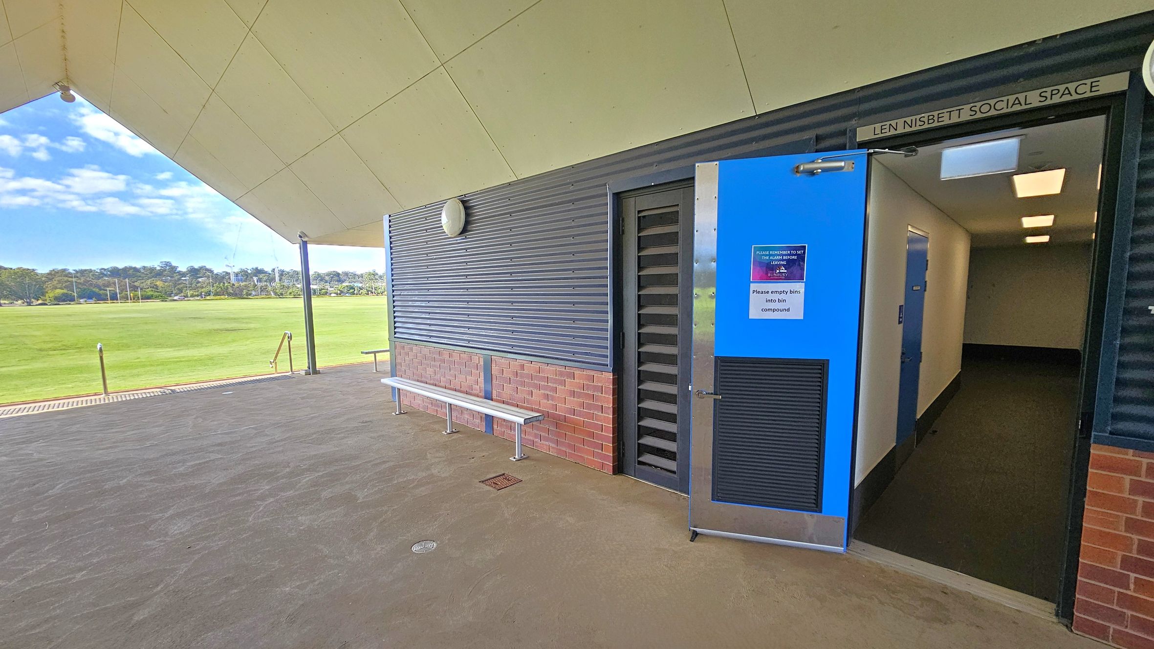 A covered outdoor area with a view of a grassy field in the background. On the right, a blue door is open, leading into a hallway with white walls and a dark floor. Above the door, a sign reads "Len Nisbett Social Space." The exterior wall is a combination of corrugated metal and brick, with a white bench attached. The ceiling is angular and light-coloured, supported by a black pole. The floor is concrete, and there are metal railings leading down to the field. A covered outdoor area with a view of a grassy field in the background. On the right, a blue door is open, leading into a hallway with white walls and a dark floor. Above the door, a sign reads "Len Nisbett Social Space." The exterior wall is a combination of corrugated metal and brick, with a white bench attached. The ceiling is angular and light-coloured, supported by a black pole. The floor is concrete, and there are metal railings leading down to the field.