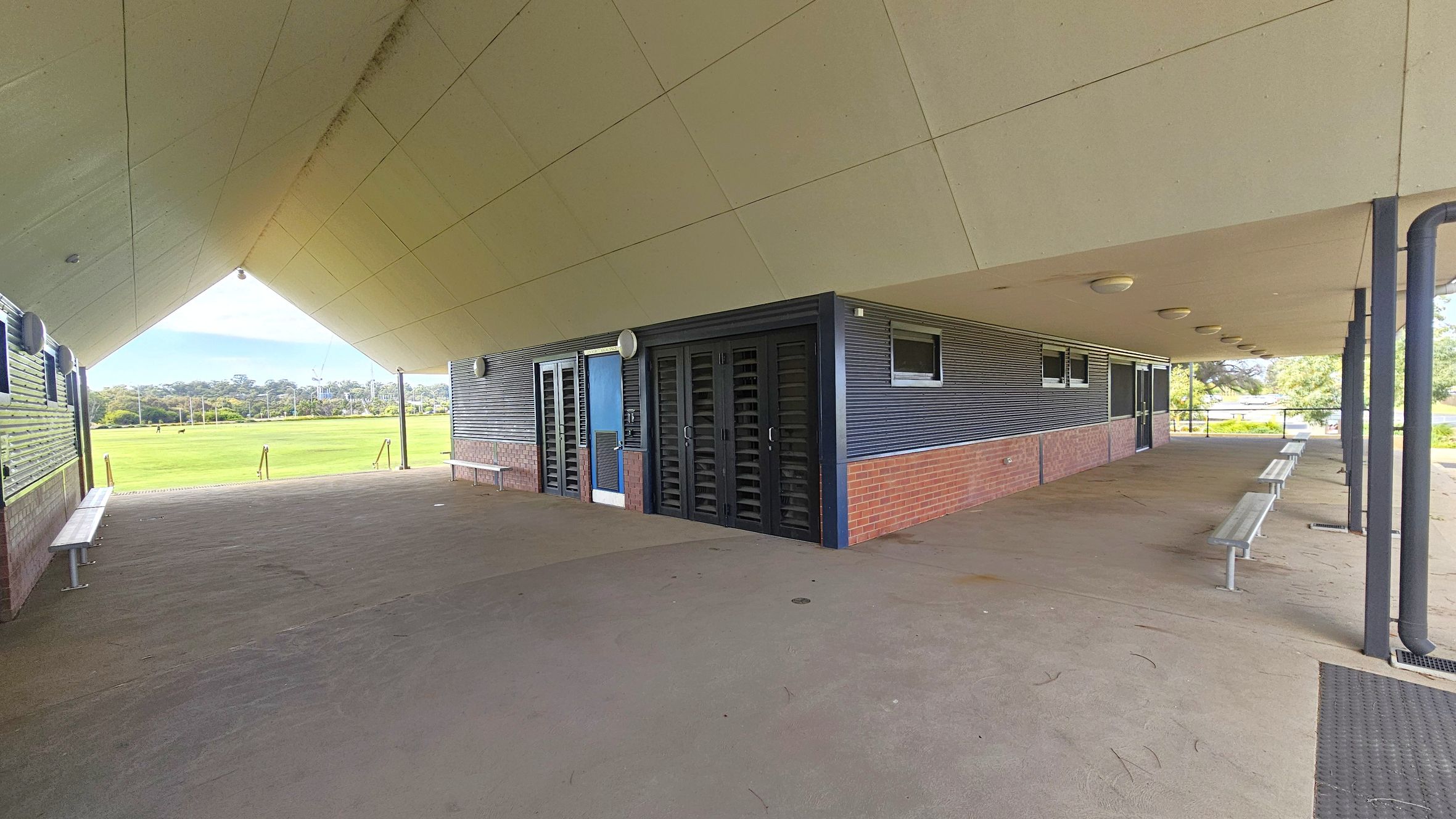 A covered outdoor area with a sloped ceiling and a concrete floor. On the right, there are metal benches and a structure with brick and corrugated metal walls, featuring doors and small windows. The left side opens to a grassy field with trees in the distance. The ceiling has several round lights, and the area is supported by metal columns. A covered outdoor area with a sloped ceiling and a concrete floor. On the right, there are metal benches and a structure with brick and corrugated metal walls, featuring doors and small windows. The left side opens to a grassy field with trees in the distance. The ceiling has several round lights, and the area is supported by metal columns.