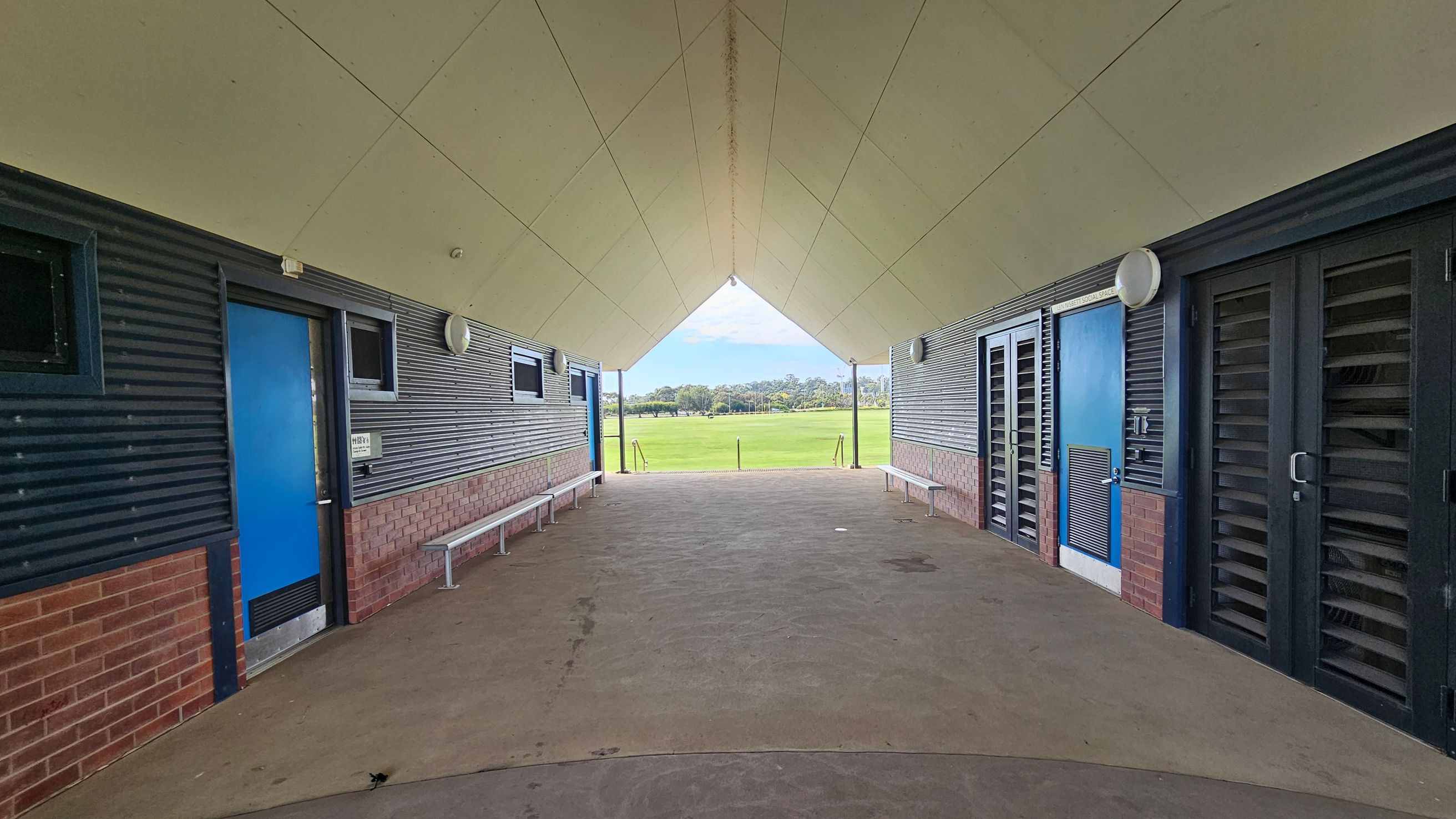 A covered walkway with a triangular ceiling leads to an open grassy field in the background. On the left, there is a blue door and a bench along a wall with corrugated metal and brick. On the right, similar walls feature a blue door and louvred panels. The floor is concrete, and the ceiling is light-coloured with a central seam. A covered walkway with a triangular ceiling leads to an open grassy field in the background. On the left, there is a blue door and a bench along a wall with corrugated metal and brick. On the right, similar walls feature a blue door and louvred panels. The floor is concrete, and the ceiling is light-coloured with a central seam.