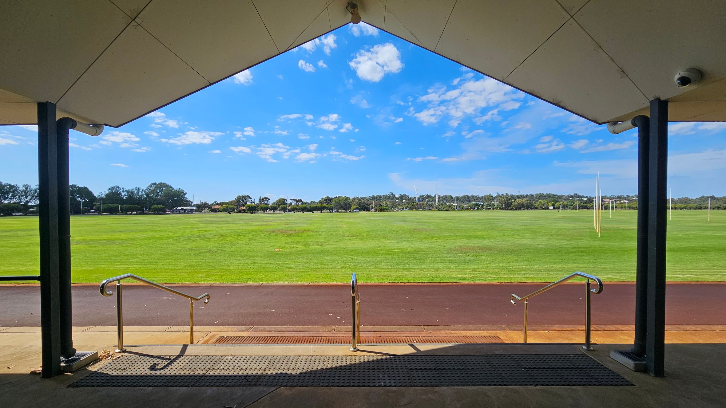 A view from under a sheltered area with a triangular roof, supported by black columns. In the foreground, metal railings lead down steps to a paved path. Beyond the path lies a large, well-maintained grassy field with several tall goalposts. The background features a line of trees and some distant structures. The ceiling of the shelter is light-coloured, and the floor is concrete with a textured mat near the steps. A view from under a sheltered area with a triangular roof, supported by black columns. In the foreground, metal railings lead down steps to a paved path. Beyond the path lies a large, well-maintained grassy field with several tall goalposts. The background features a line of trees and some distant structures. The ceiling of the shelter is light-coloured, and the floor is concrete with a textured mat near the steps.