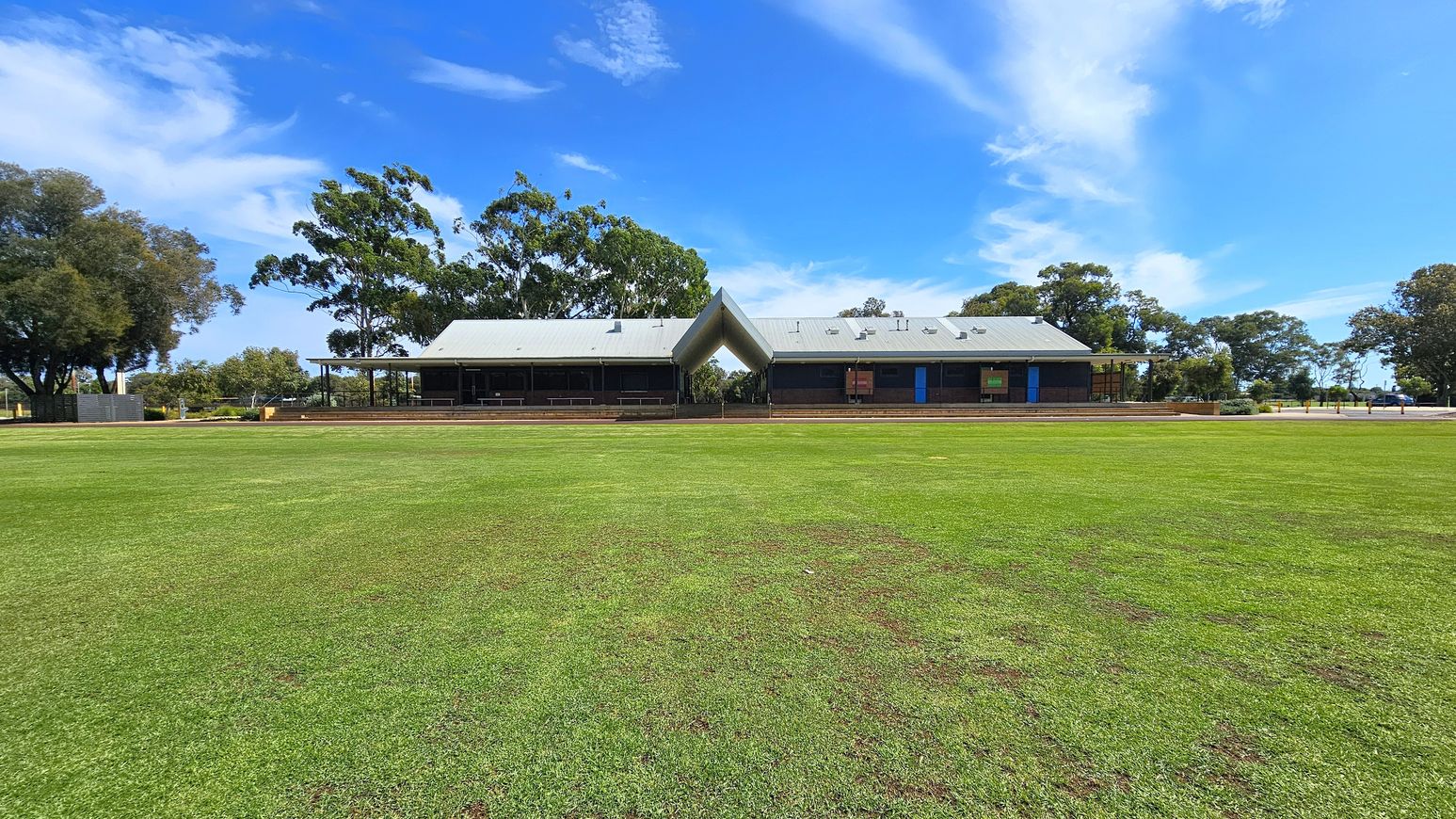 A large, open grassy field in the foreground leads to a modern building in the centre. The building has a distinctive V-shaped roof and is flanked by trees on both sides. The structure features a covered veranda and large windows. The sky is clear with some clouds, and the area is surrounded by more trees, creating a natural backdrop. A large, open grassy field in the foreground leads to a modern building in the centre. The building has a distinctive V-shaped roof and is flanked by trees on both sides. The structure features a covered veranda and large windows. The sky is clear with some clouds, and the area is surrounded by more trees, creating a natural backdrop.