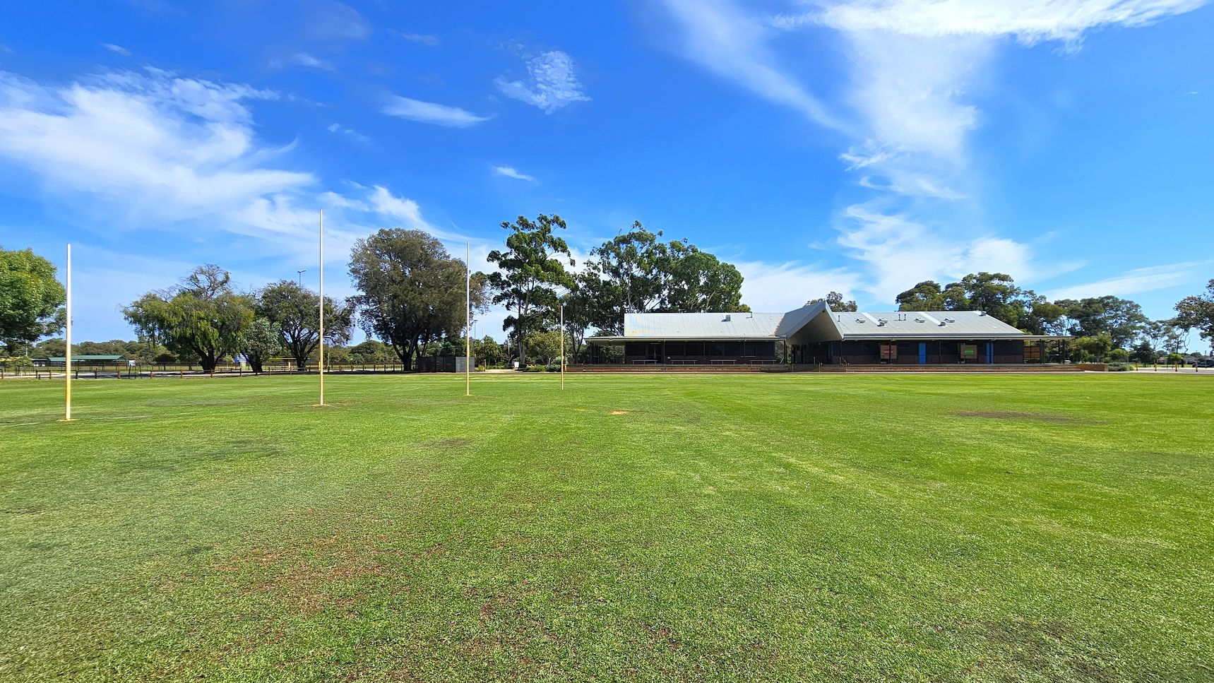 A grassy sports field with four tall goalposts in the centre. In the background, there is a building with a grey roof and a veranda, surrounded by several trees. The field is expansive, with a well-maintained green surface. The sky is clear with some clouds. A grassy sports field with four tall goalposts in the centre. In the background, there is a building with a grey roof and a veranda, surrounded by several trees. The field is expansive, with a well-maintained green surface. The sky is clear with some clouds.