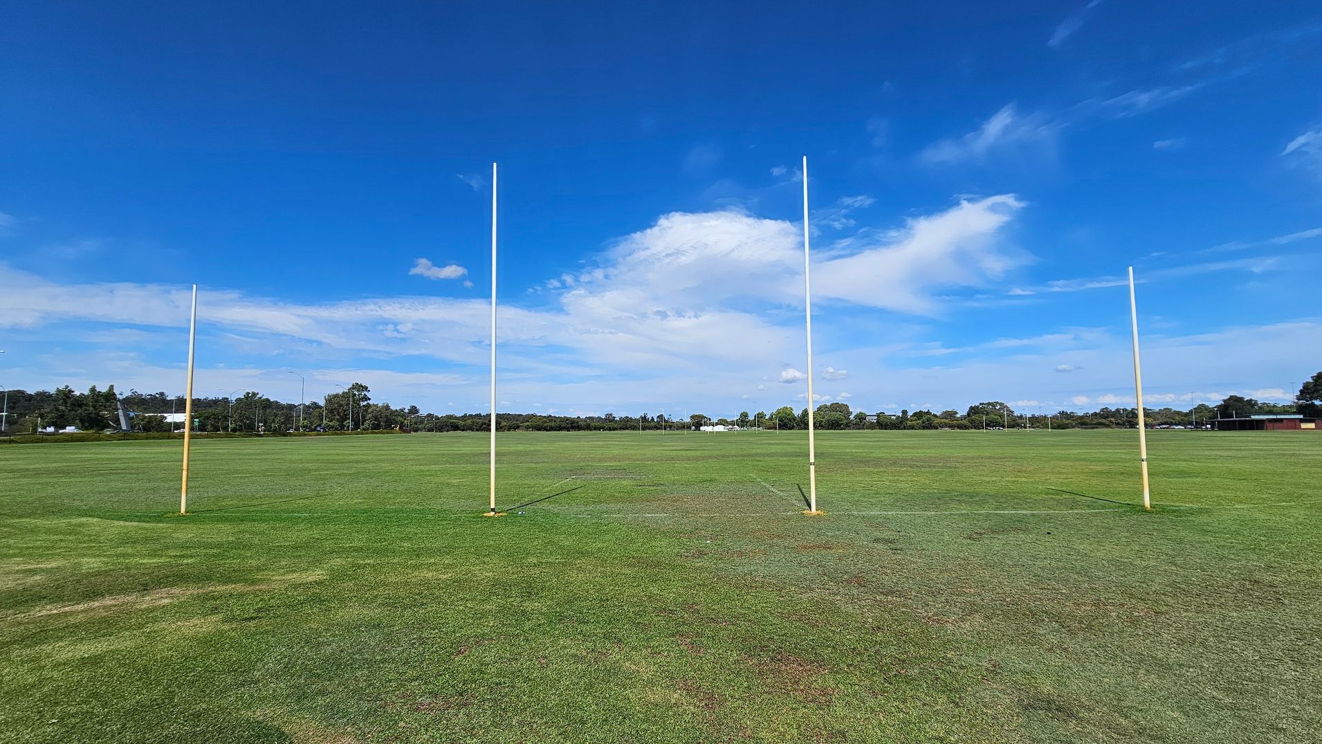 Four tall goalposts stand on a large, grassy field. The grass is a mix of green and brown patches. In the background, there are trees and some low buildings. The sky is clear with a few clouds. Four tall goalposts stand on a large, grassy field. The grass is a mix of green and brown patches. In the background, there are trees and some low buildings. The sky is clear with a few clouds.