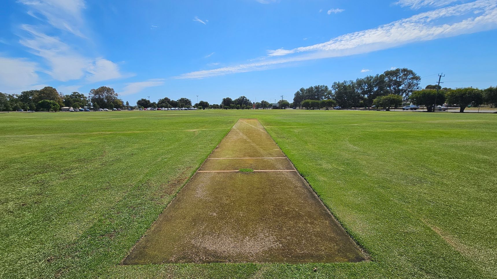 A cricket pitch is in the centre of a large grassy field. The pitch is a rectangular strip with a worn, brown surface. Surrounding the pitch is a well-maintained green outfield. In the background, there are trees and a line of parked cars along a road. The sky is blue with some clouds. A cricket pitch is in the centre of a large grassy field. The pitch is a rectangular strip with a worn, brown surface. Surrounding the pitch is a well-maintained green outfield. In the background, there are trees and a line of parked cars along a road. The sky is blue with some clouds.