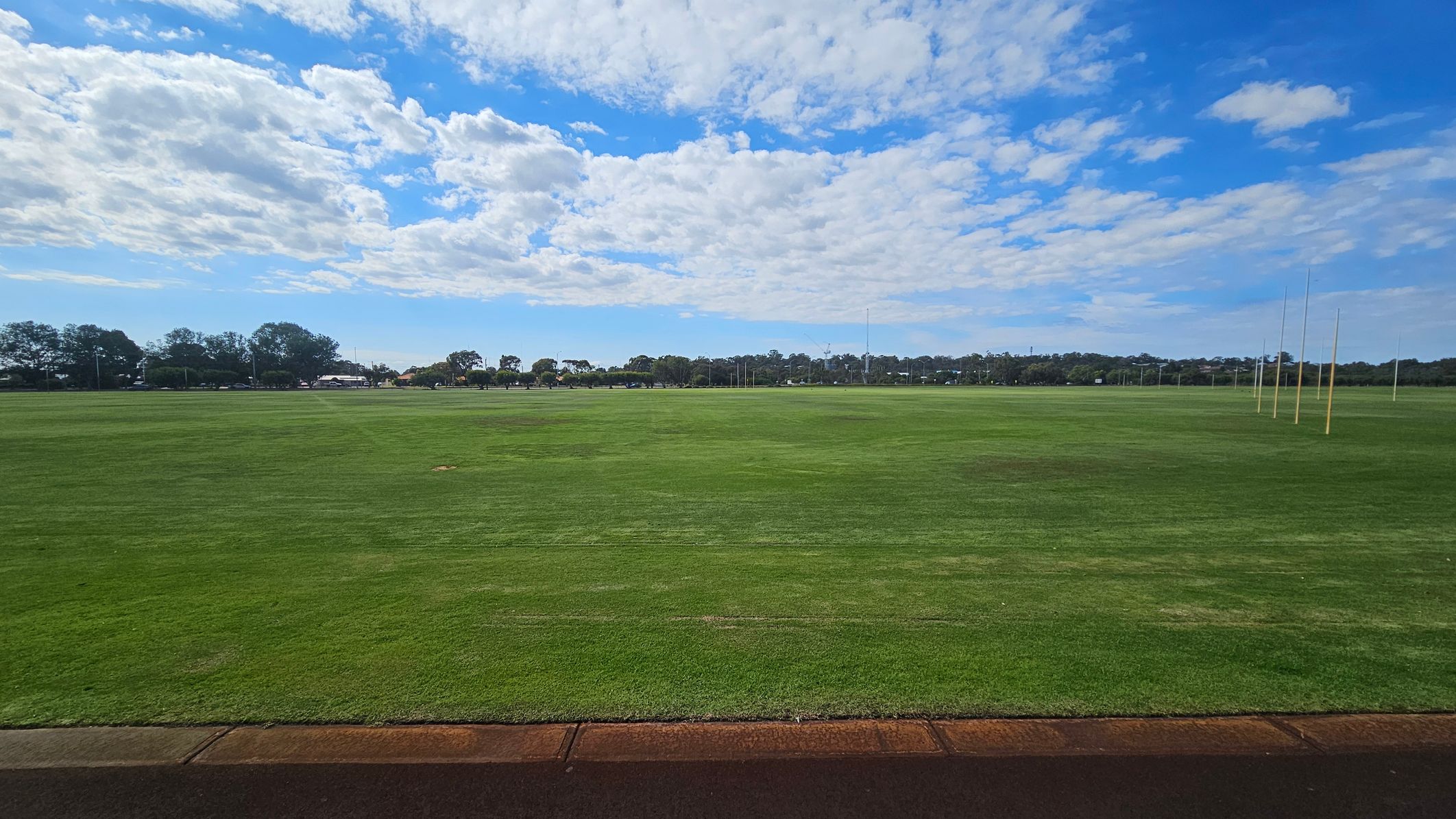 A wide, open grassy field stretches across the image, with a few patches of brown. On the right, several tall goalposts are visible. In the background, a line of trees and some distant structures can be seen. The foreground features a narrow strip of pavement. The sky is partly cloudy with patches of blue. A wide, open grassy field stretches across the image, with a few patches of brown. On the right, several tall goalposts are visible. In the background, a line of trees and some distant structures can be seen. The foreground features a narrow strip of pavement. The sky is partly cloudy with patches of blue.