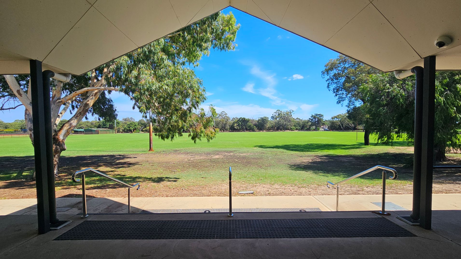 A view from under a sheltered area with a triangular ceiling, supported by black columns. In the foreground, there are steps with metal railings leading down to a grassy field. The field is expansive and bordered by trees on both sides. A large tree is visible on the left, and another on the right, with more trees in the distance. The ground near the steps is a mix of grass and bare patches. A view from under a sheltered area with a triangular ceiling, supported by black columns. In the foreground, there are steps with metal railings leading down to a grassy field. The field is expansive and bordered by trees on both sides. A large tree is visible on the left, and another on the right, with more trees in the distance. The ground near the steps is a mix of grass and bare patches.