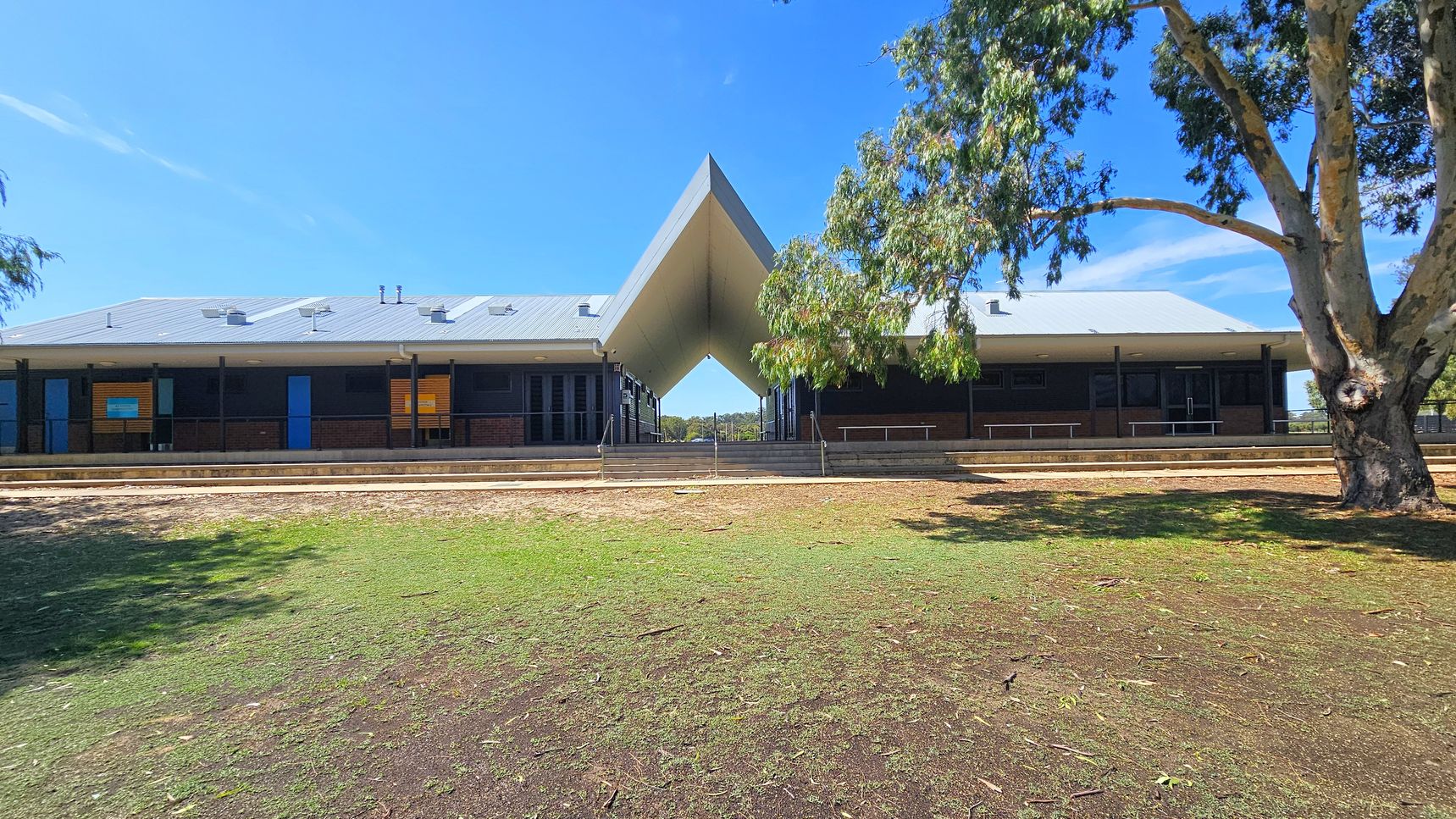 A building with a distinctive V-shaped roof structure in the centre. The building has a grey roof and dark walls with several blue doors and windows. In the foreground, there is a grassy area with patches of dirt and a large tree on the right. The background shows a clear blue sky. The building is elevated with steps leading up to it, and there are railings along the front. A building with a distinctive V-shaped roof structure in the centre. The building has a grey roof and dark walls with several blue doors and windows. In the foreground, there is a grassy area with patches of dirt and a large tree on the right. The background shows a clear blue sky. The building is elevated with steps leading up to it, and there are railings along the front.