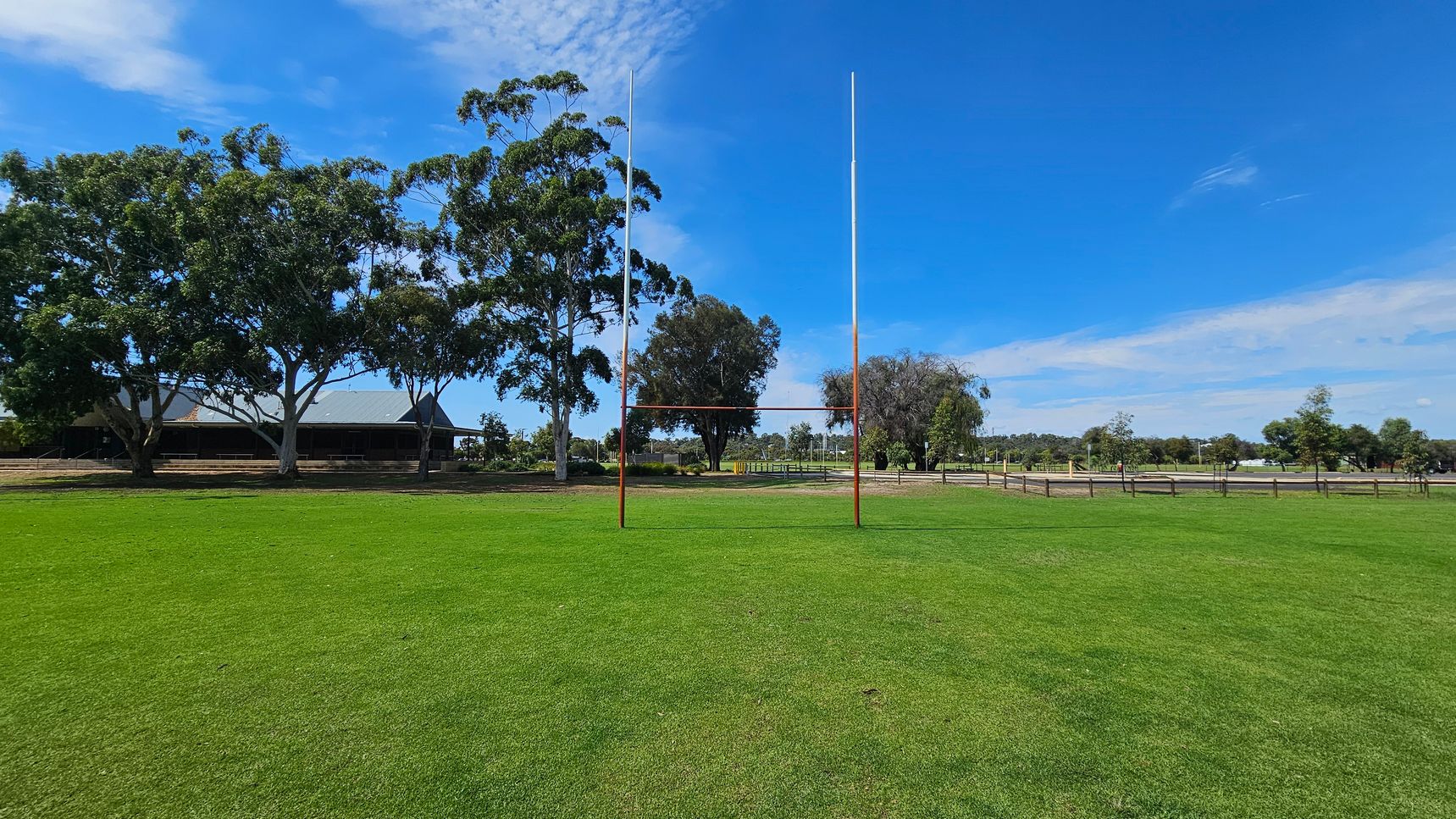 A rugby field with bright green grass is in the foreground. In the centre, there are rugby posts. To the left, large trees provide shade, and a building with a dark roof is partially visible. On the right, more trees and a wooden fence line the edge of the field. The background features a mix of trees and open space. A rugby field with bright green grass is in the foreground. In the centre, there are rugby posts. To the left, large trees provide shade, and a building with a dark roof is partially visible. On the right, more trees and a wooden fence line the edge of the field. The background features a mix of trees and open space.