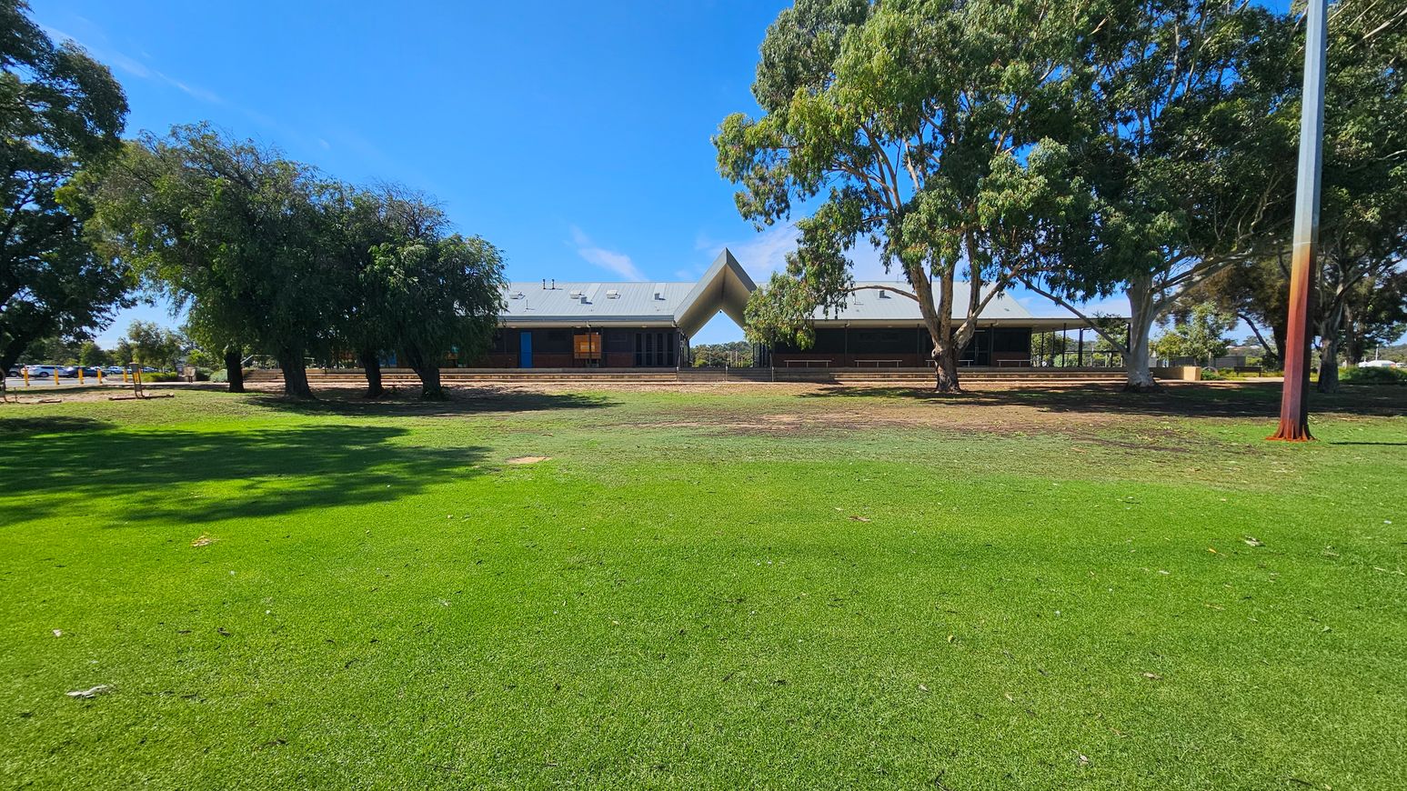 A grassy field in the foreground leads to a building with a distinctive triangular roof structure in the centre. Trees with dense foliage are on both sides, providing shade. The building has a grey roof with several vents and a blue door visible on the left side. A tall pole stands on the right side of the image. The ground near the building is a mix of grass and bare earth. A grassy field in the foreground leads to a building with a distinctive triangular roof structure in the centre. Trees with dense foliage are on both sides, providing shade. The building has a grey roof with several vents and a blue door visible on the left side. A tall pole stands on the right side of the image. The ground near the building is a mix of grass and bare earth.
