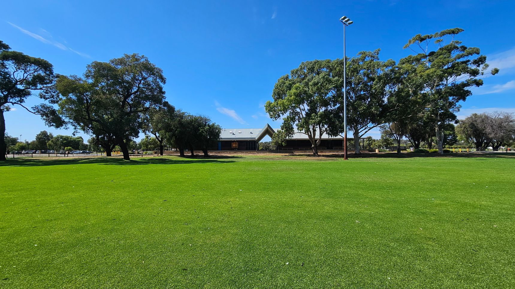 A wide expanse of green grass covers the foreground. In the background, there are several large trees with dense foliage. A building with a peaked roof is partially visible behind the trees. A tall light pole stands to the right of the building. The sky is clear and blue. A wide expanse of green grass covers the foreground. In the background, there are several large trees with dense foliage. A building with a peaked roof is partially visible behind the trees. A tall light pole stands to the right of the building. The sky is clear and blue.