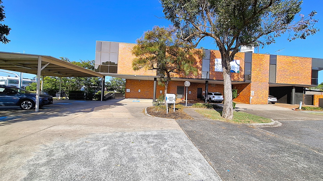 A car park with a covered area on the left, housing several parked cars. In the middle, a tree stands in front of a two-storey brick building with large windows. The building has a sign and a circular driveway leading to the entrance. On the right, more parking spaces are visible, with additional cars parked. The ground is a mix of concrete and asphalt, and the area is surrounded by greenery. A car park with a covered area on the left, housing several parked cars. In the middle, a tree stands in front of a two-storey brick building with large windows. The building has a sign and a circular driveway leading to the entrance. On the right, more parking spaces are visible, with additional cars parked. The ground is a mix of concrete and asphalt, and the area is surrounded by greenery.