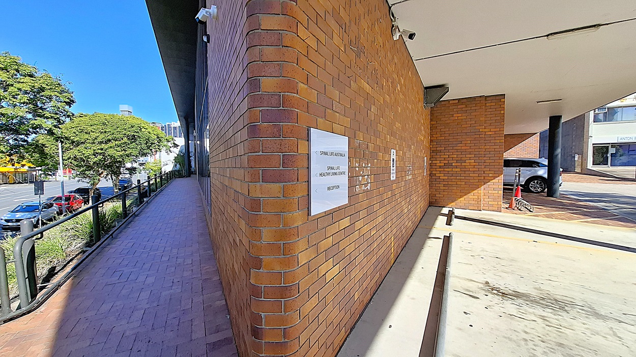 On the left, a brick walkway with a black metal railing runs alongside a row of trees and parked cars. In the middle, a brick wall with signs and a security camera. On the right, a covered parking area with a concrete floor, a black column, and a parked car. The ceiling is white with light fixtures and seams. On the left, a brick walkway with a black metal railing runs alongside a row of trees and parked cars. In the middle, a brick wall with signs and a security camera. On the right, a covered parking area with a concrete floor, a black column, and a parked car. The ceiling is white with light fixtures and seams.