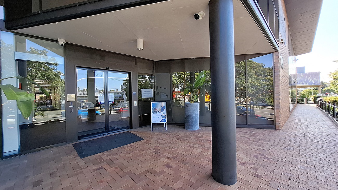 On the left, large glass windows reflect the surrounding trees and buildings. In the middle, a glass entrance door with a black frame, flanked by a signboard and a large potted plant. On the right, a black column supports the overhang, with more glass windows and a brick walkway extending along the building. The floor is made of red bricks, and the ceiling is white with light fixtures and a security camera. On the left, large glass windows reflect the surrounding trees and buildings. In the middle, a glass entrance door with a black frame, flanked by a signboard and a large potted plant. On the right, a black column supports the overhang, with more glass windows and a brick walkway extending along the building. The floor is made of red bricks, and the ceiling is white with light fixtures and a security camera.