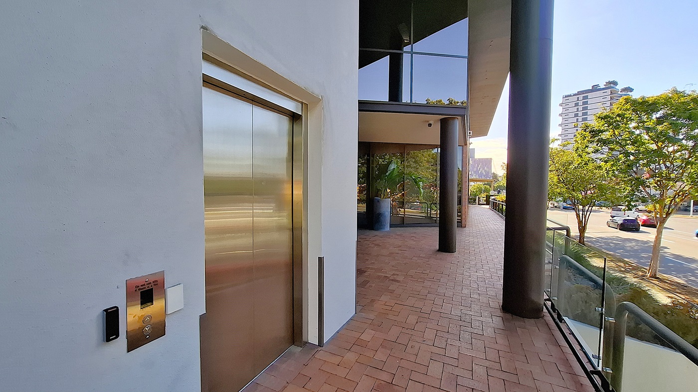 On the left, a stainless steel lift door set in a white wall with a control panel beside it. In the middle, a large black column supports an overhang, with glass windows and a potted plant visible. On the right, a brick walkway bordered by a glass railing, leading to a street with trees and parked cars. The floor is red brick, and the ceiling is white with recessed lighting. On the left, a stainless steel lift door set in a white wall with a control panel beside it. In the middle, a large black column supports an overhang, with glass windows and a potted plant visible. On the right, a brick walkway bordered by a glass railing, leading to a street with trees and parked cars. The floor is red brick, and the ceiling is white with recessed lighting.