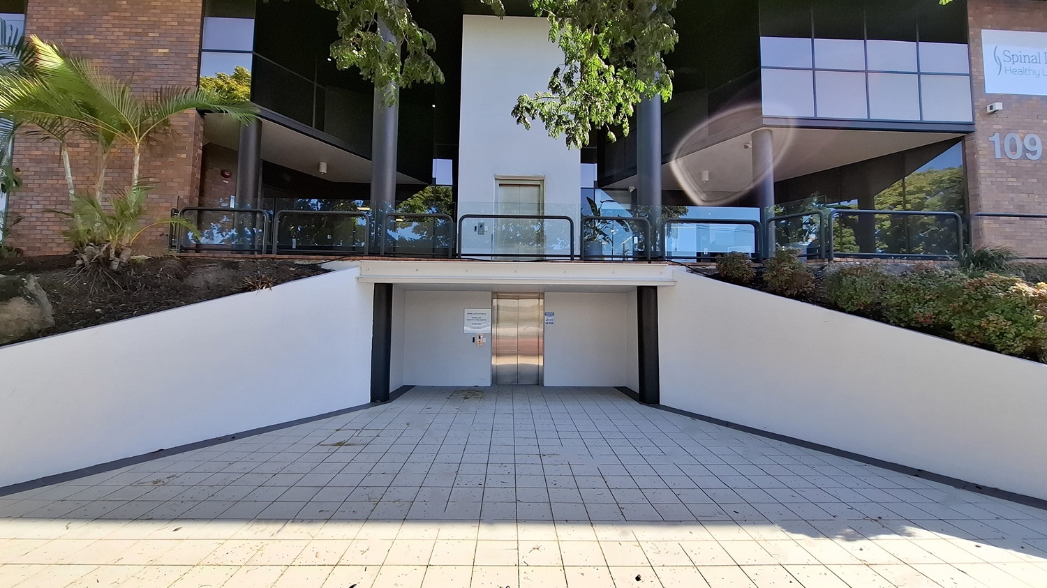 On the left, a brick wall with a small garden area containing palm trees and shrubs. In the middle, a stainless steel lift door set into a white wall, with a tiled ramp leading up to it. On the right, another brick wall with a sign and more greenery. The floor is tiled with light-coloured square tiles, and the ceiling above the lift area is white with recessed lighting. On the left, a brick wall with a small garden area containing palm trees and shrubs. In the middle, a stainless steel lift door set into a white wall, with a tiled ramp leading up to it. On the right, another brick wall with a sign and more greenery. The floor is tiled with light-coloured square tiles, and the ceiling above the lift area is white with recessed lighting.