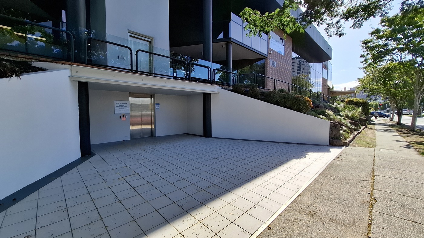 On the left, a white wall with a black column and a glass railing above. In the middle, a stainless steel lift door with a control panel and signs on either side. On the right, a sloped garden area with shrubs and a black railing, leading to a brick building with large windows. The floor is tiled with light-coloured square tiles, and the ceiling above the lift area is white. On the left, a white wall with a black column and a glass railing above. In the middle, a stainless steel lift door with a control panel and signs on either side. On the right, a sloped garden area with shrubs and a black railing, leading to a brick building with large windows. The floor is tiled with light-coloured square tiles, and the ceiling above the lift area is white.