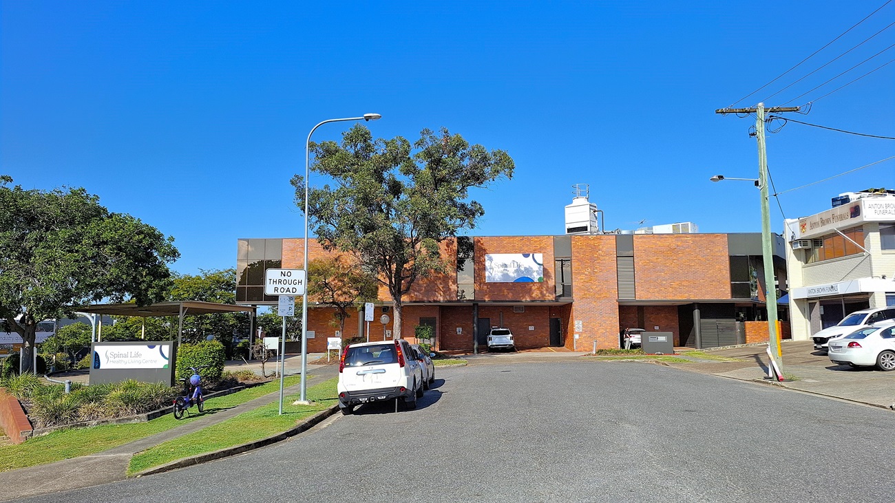 On the left, a covered walkway with a sign reading "Spinal Life" and a person on a bicycle. In the middle, a two-storey brick building with a large tree in front, a "No Through Road" sign, and several parked cars. On the right, a utility pole with wires and a white building with signage. The ground is paved with asphalt, and there is a grassy area with shrubs near the walkway. On the left, a covered walkway with a sign reading "Spinal Life" and a person on a bicycle. In the middle, a two-storey brick building with a large tree in front, a "No Through Road" sign, and several parked cars. On the right, a utility pole with wires and a white building with signage. The ground is paved with asphalt, and there is a grassy area with shrubs near the walkway.