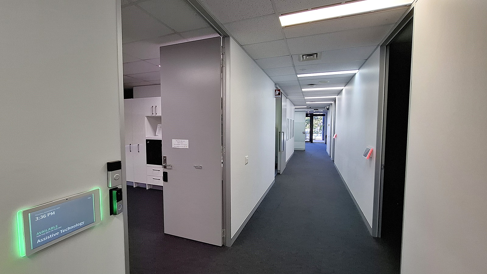 On the left, a large television screen mounted on the wall above a display table with a model labelled "Spinal Life Healthy Living Centre" and a potted plant beside it. In the middle, a long hallway with white walls, a grey carpet, and fluorescent ceiling lights, leading to a glass door at the end. On the right, a white reception desk with a computer monitor and informational posters on the wall. The ceiling is white with recessed lighting. On the left, a large television screen mounted on the wall above a display table with a model labelled "Spinal Life Healthy Living Centre" and a potted plant beside it. In the middle, a long hallway with white walls, a grey carpet, and fluorescent ceiling lights, leading to a glass door at the end. On the right, a white reception desk with a computer monitor and informational posters on the wall. The ceiling is white with recessed lighting.