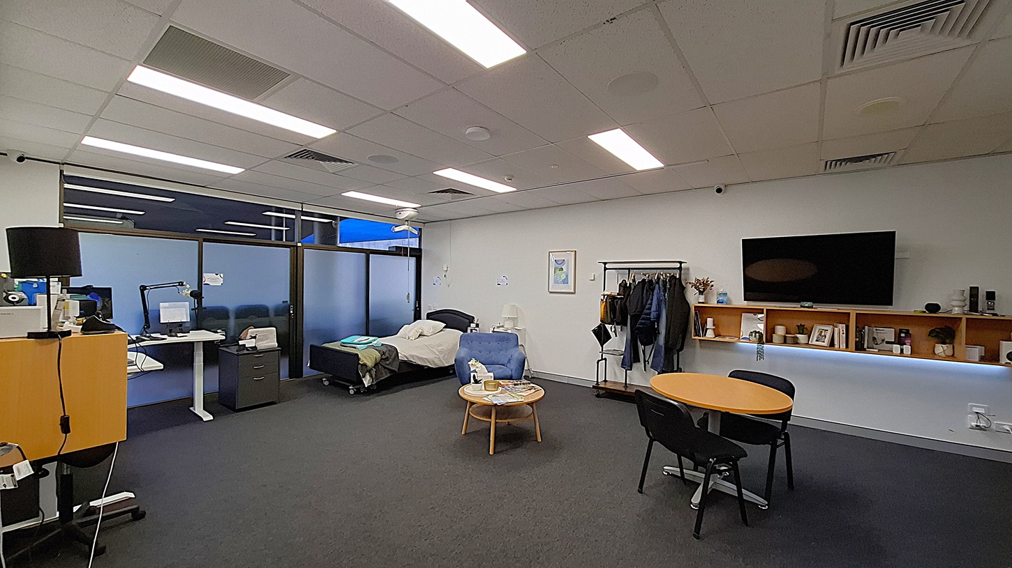 On the left, a desk with a computer monitor, a black lamp, and a filing cabinet. In the middle, a bed with white sheets and a blue blanket, a small table with a tissue box and a water bottle, and a blue armchair. On the right, a wall-mounted television above wooden shelves holding various items, and a coat rack with jackets. The floor is dark grey carpet, and the ceiling has white panels with fluorescent lighting. On the left, a desk with a computer monitor, a black lamp, and a filing cabinet. In the middle, a bed with white sheets and a blue blanket, a small table with a tissue box and a water bottle, and a blue armchair. On the right, a wall-mounted television above wooden shelves holding various items, and a coat rack with jackets. The floor is dark grey carpet, and the ceiling has white panels with fluorescent lighting.