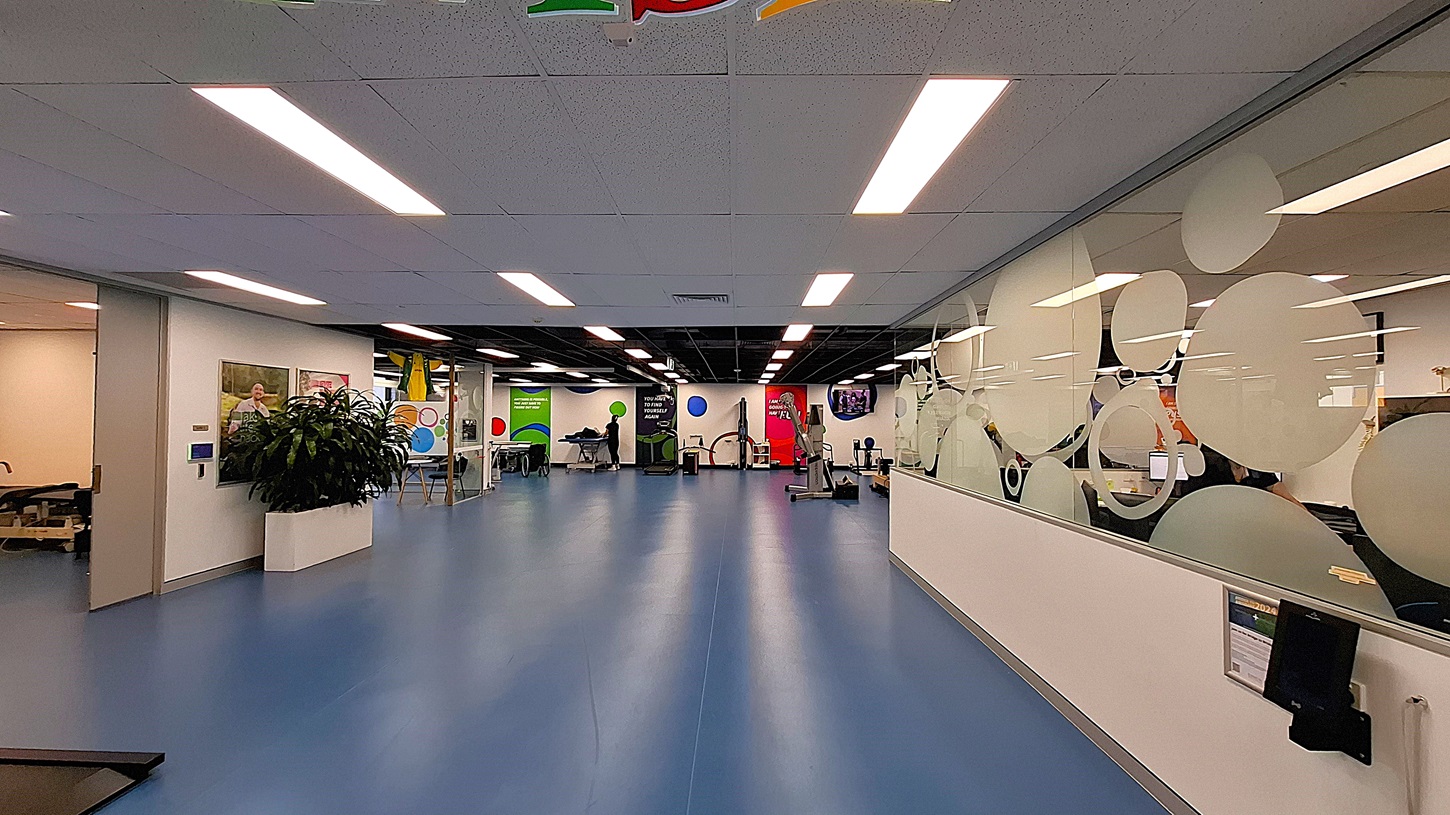 On the left, an open area with blue flooring, exercise equipment, and colourful wall decorations. In the middle, a glass partition with frosted circular designs and a "Finish Line" banner hanging above. On the right, a hallway with dark carpet, a white shelving unit holding various items and a potted plant, and a black bin. The ceiling has white panels with recessed lighting and air vents. On the left, an open area with blue flooring, exercise equipment, and colourful wall decorations. In the middle, a glass partition with frosted circular designs and a "Finish Line" banner hanging above. On the right, a hallway with dark carpet, a white shelving unit holding various items and a potted plant, and a black bin. The ceiling has white panels with recessed lighting and air vents.