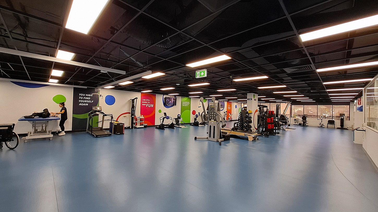 On the left, a person working on a black exercise table next to a wall with motivational text and colourful circular designs. In the middle, various exercise machines including treadmills and stationary bikes, with bright wall graphics. On the right, more exercise equipment, weight racks, and a glass partition with a view of an office area. The floor is blue, and the ceiling is black with exposed structures and recessed lighting. On the left, a person working on a black exercise table next to a wall with motivational text and colourful circular designs. In the middle, various exercise machines including treadmills and stationary bikes, with bright wall graphics. On the right, more exercise equipment, weight racks, and a glass partition with a view of an office area. The floor is blue, and the ceiling is black with exposed structures and recessed lighting.