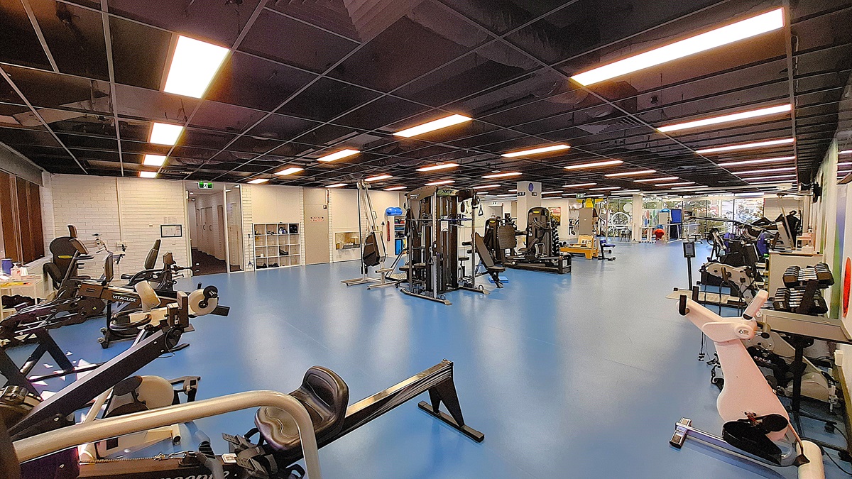 On the left, various exercise machines including stationary bikes and rowing machines, positioned near a white brick wall with a small window. In the middle, a spacious area with blue flooring and multiple weight and resistance training machines. On the right, more exercise equipment including treadmills and weight racks, with a large window in the background. The ceiling is black with exposed structures and evenly spaced fluorescent lighting. On the left, various exercise machines including stationary bikes and rowing machines, positioned near a white brick wall with a small window. In the middle, a spacious area with blue flooring and multiple weight and resistance training machines. On the right, more exercise equipment including treadmills and weight racks, with a large window in the background. The ceiling is black with exposed structures and evenly spaced fluorescent lighting.