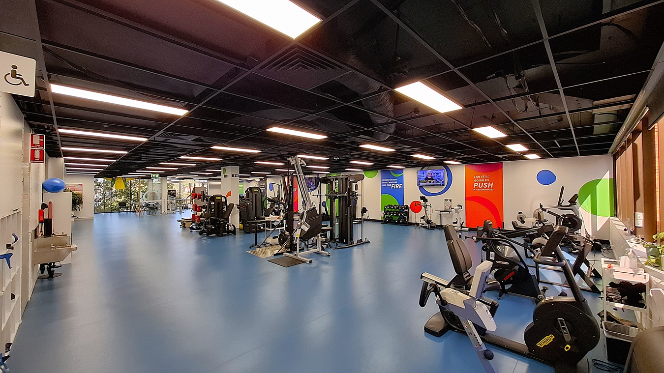 On the left, a white wall with a sink, shelves holding exercise balls, and a fire extinguisher. In the middle, a spacious area with blue flooring, various exercise machines including weight and resistance training equipment, and colourful motivational banners on the walls. On the right, more exercise equipment including stationary bikes and treadmills, with large windows letting in natural light. The ceiling is black with exposed structures and evenly spaced fluorescent lighting. On the left, a white wall with a sink, shelves holding exercise balls, and a fire extinguisher. In the middle, a spacious area with blue flooring, various exercise machines including weight and resistance training equipment, and colourful motivational banners on the walls. On the right, more exercise equipment including stationary bikes and treadmills, with large windows letting in natural light. The ceiling is black with exposed structures and evenly spaced fluorescent lighting.