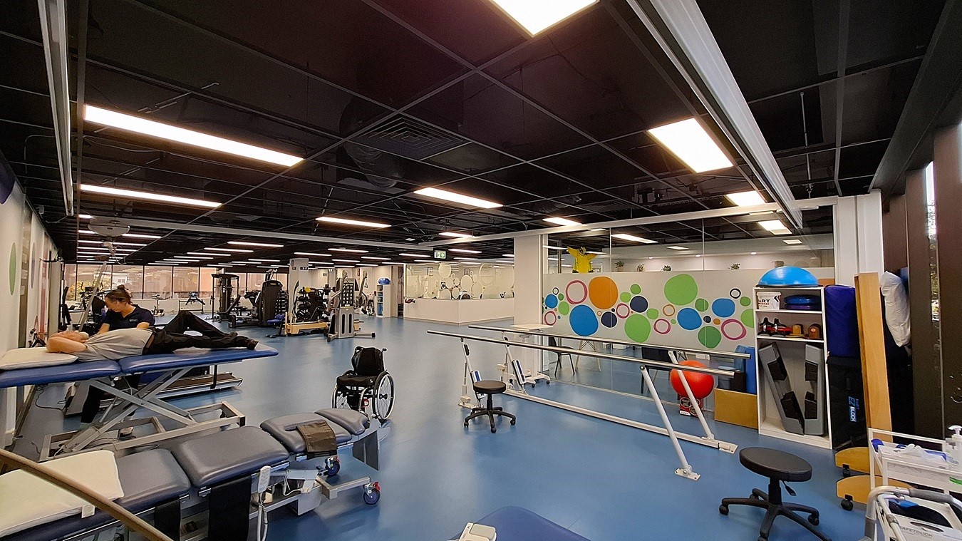 On the left, a person working on a blue treatment table next to a wall with colourful circular designs. In the middle, a spacious area with blue flooring, various exercise machines, and a wheelchair. On the right, a white shelving unit holding exercise balls and equipment, and a parallel bar setup for rehabilitation. The ceiling is black with exposed structures and evenly spaced fluorescent lighting. On the left, a person working on a blue treatment table next to a wall with colourful circular designs. In the middle, a spacious area with blue flooring, various exercise machines, and a wheelchair. On the right, a white shelving unit holding exercise balls and equipment, and a parallel bar setup for rehabilitation. The ceiling is black with exposed structures and evenly spaced fluorescent lighting.