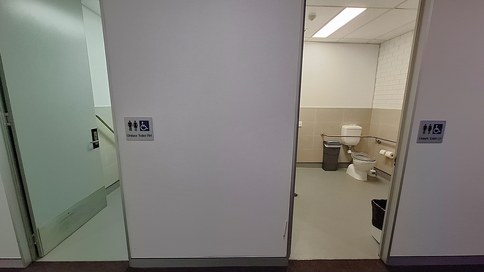 On the left, an open door leading to a stairwell with a handrail and a sign indicating a unisex accessible toilet. In the middle, a white wall separating two doorways. On the right, an open door revealing a bathroom with a toilet, grab bars, a waste bin, and a toilet paper holder. The floor is grey, and the ceiling has white panels with fluorescent lighting. On the left, an open door leading to a stairwell with a handrail and a sign indicating a unisex accessible toilet. In the middle, a white wall separating two doorways. On the right, an open door revealing a bathroom with a toilet, grab bars, a waste bin, and a toilet paper holder. The floor is grey, and the ceiling has white panels with fluorescent lighting.