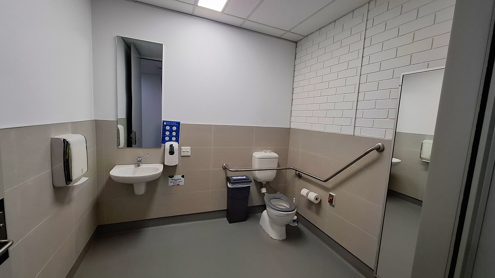 On the left, a white sink with a mirror above it, a soap dispenser, and a paper towel dispenser. In the middle, a grey waste bin and a white toilet with a grab bar on the right side. On the right, a large mirror reflecting the sink area and a toilet paper holder mounted on the wall. The floor is grey, and the ceiling has white panels with fluorescent lighting. The walls are half-tiled in beige with the upper half painted white, and one wall has white brickwork. On the left, a white sink with a mirror above it, a soap dispenser, and a paper towel dispenser. In the middle, a grey waste bin and a white toilet with a grab bar on the right side. On the right, a large mirror reflecting the sink area and a toilet paper holder mounted on the wall. The floor is grey, and the ceiling has white panels with fluorescent lighting. The walls are half-tiled in beige with the upper half painted white, and one wall has white brickwork.