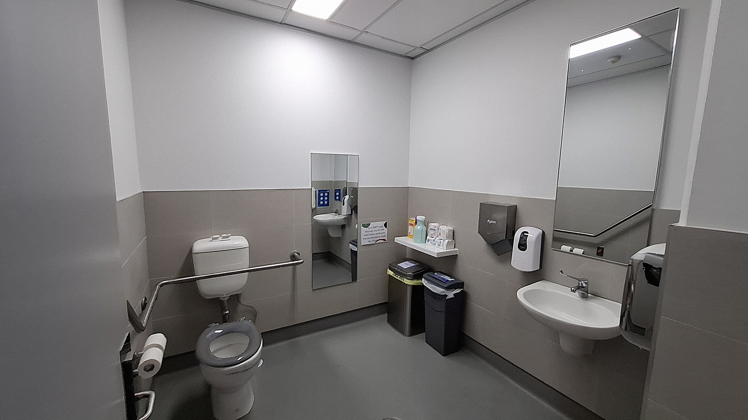 On the left, a white toilet with a grey seat, a grab bar, and a toilet paper holder. In the middle, a wall-mounted mirror above a shelf holding cleaning supplies, with two bins below. On the right, a white sink with a soap dispenser, a paper towel dispenser, and a large mirror above. The floor is grey, and the ceiling has white panels with fluorescent lighting. The walls are half-tiled in beige with the upper half painted white. On the left, a white toilet with a grey seat, a grab bar, and a toilet paper holder. In the middle, a wall-mounted mirror above a shelf holding cleaning supplies, with two bins below. On the right, a white sink with a soap dispenser, a paper towel dispenser, and a large mirror above. The floor is grey, and the ceiling has white panels with fluorescent lighting. The walls are half-tiled in beige with the upper half painted white.
