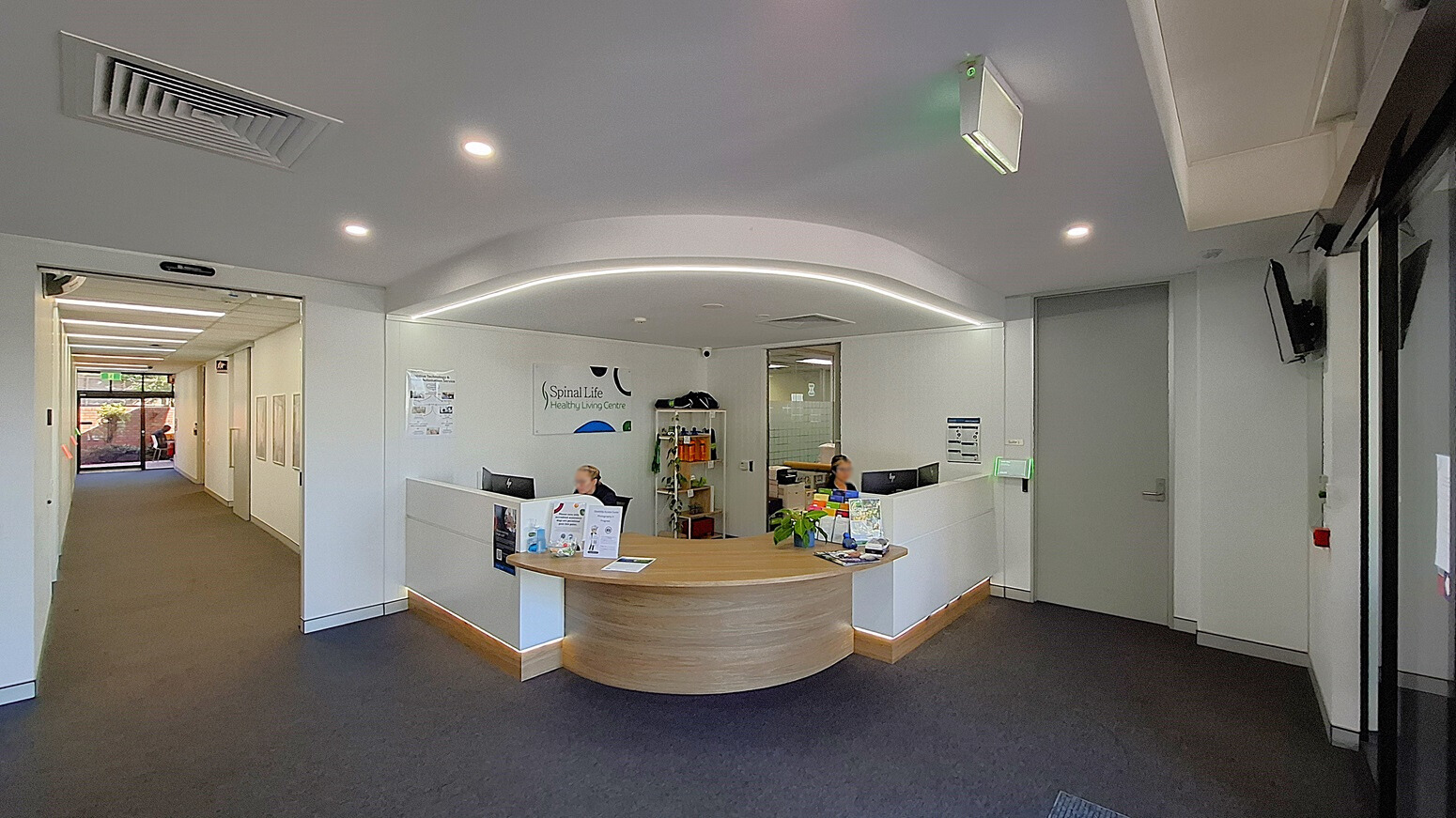 On the left, a large potted plant with broad leaves stands next to a glass wall. In the middle, a glass entrance door with a black frame leads to a reception area with a curved wooden desk and a person seated behind it. On the right, a signboard with business hours and another potted plant. The floor is covered with red bricks outside and dark carpet inside. The ceiling is white with recessed lighting and a security camera above the entrance. On the left, a large potted plant with broad leaves stands next to a glass wall. In the middle, a glass entrance door with a black frame leads to a reception area with a curved wooden desk and a person seated behind it. On the right, a signboard with business hours and another potted plant. The floor is covered with red bricks outside and dark carpet inside. The ceiling is white with recessed lighting and a security camera above the entrance.