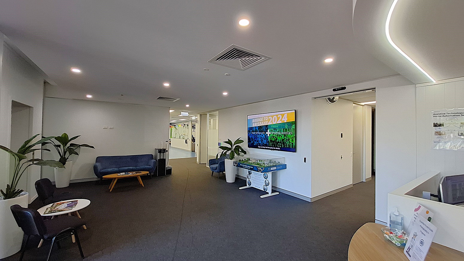 On the left, two black chairs and a small round table with magazines, next to a large potted plant. In the middle, a blue sofa with a wooden coffee table in front, and a hallway leading to another area. On the right, a wall-mounted TV displaying "Bridge to Brisbane 2024," a foosball table, and another potted plant. The floor is covered with dark carpet, and the ceiling is white with recessed lighting and air vents. On the left, two black chairs and a small round table with magazines, next to a large potted plant. In the middle, a blue sofa with a wooden coffee table in front, and a hallway leading to another area. On the right, a wall-mounted TV displaying "Bridge to Brisbane 2024," a foosball table, and another potted plant. The floor is covered with dark carpet, and the ceiling is white with recessed lighting and air vents.