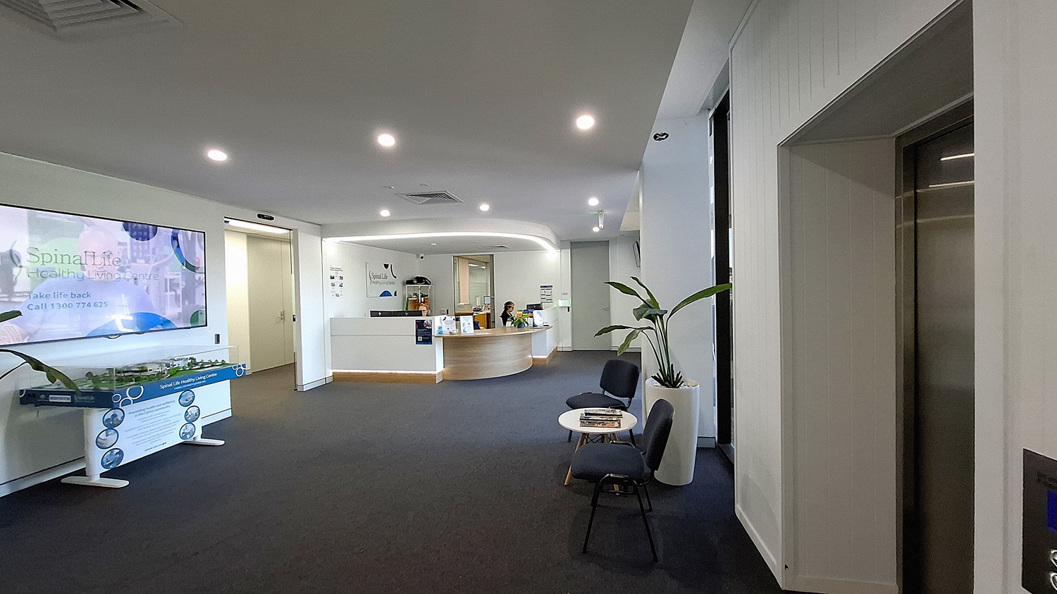 On the left, a large wall-mounted TV displaying "Spinal Life Healthy Living Centre" above a model display table. In the middle, a curved reception desk with two people seated behind it, computers, and informational brochures. On the right, a lift with stainless steel doors, a potted plant, and two black chairs with a small round table holding magazines. The floor is covered with dark carpet, and the ceiling is white with recessed lighting and air vents. On the left, a large wall-mounted TV displaying "Spinal Life Healthy Living Centre" above a model display table. In the middle, a curved reception desk with two people seated behind it, computers, and informational brochures. On the right, a lift with stainless steel doors, a potted plant, and two black chairs with a small round table holding magazines. The floor is covered with dark carpet, and the ceiling is white with recessed lighting and air vents.