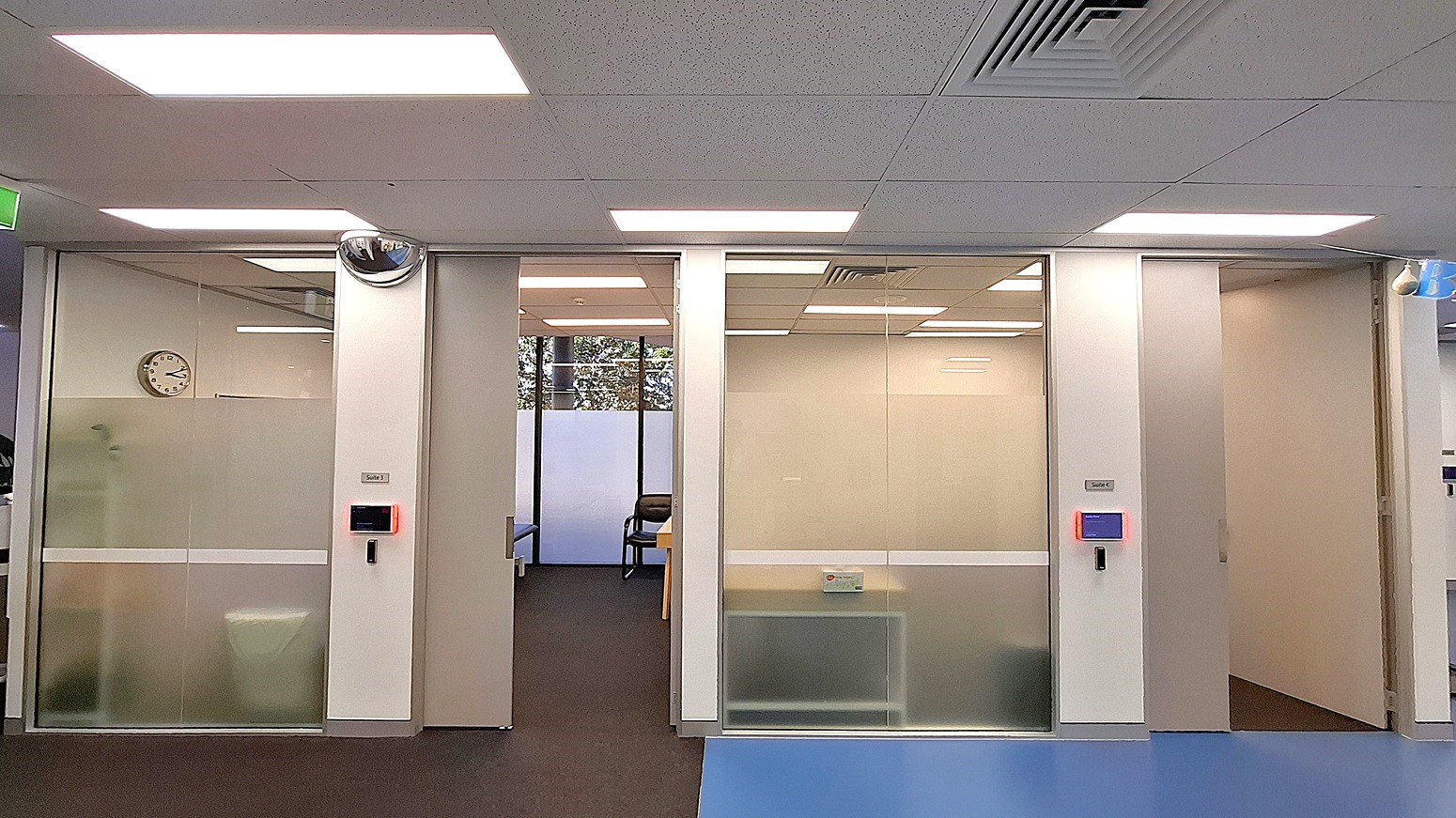 On the left, a frosted glass door with a wall-mounted clock inside the room. In the middle, an open doorway leading to a room with a chair and a table, and a window with frosted lower panels. On the right, another frosted glass door with a blue-lit access panel and a partially visible hallway. The floor is a mix of dark carpet and blue vinyl, and the ceiling is white with recessed lighting and air vents. On the left, a frosted glass door with a wall-mounted clock inside the room. In the middle, an open doorway leading to a room with a chair and a table, and a window with frosted lower panels. On the right, another frosted glass door with a blue-lit access panel and a partially visible hallway. The floor is a mix of dark carpet and blue vinyl, and the ceiling is white with recessed lighting and air vents.